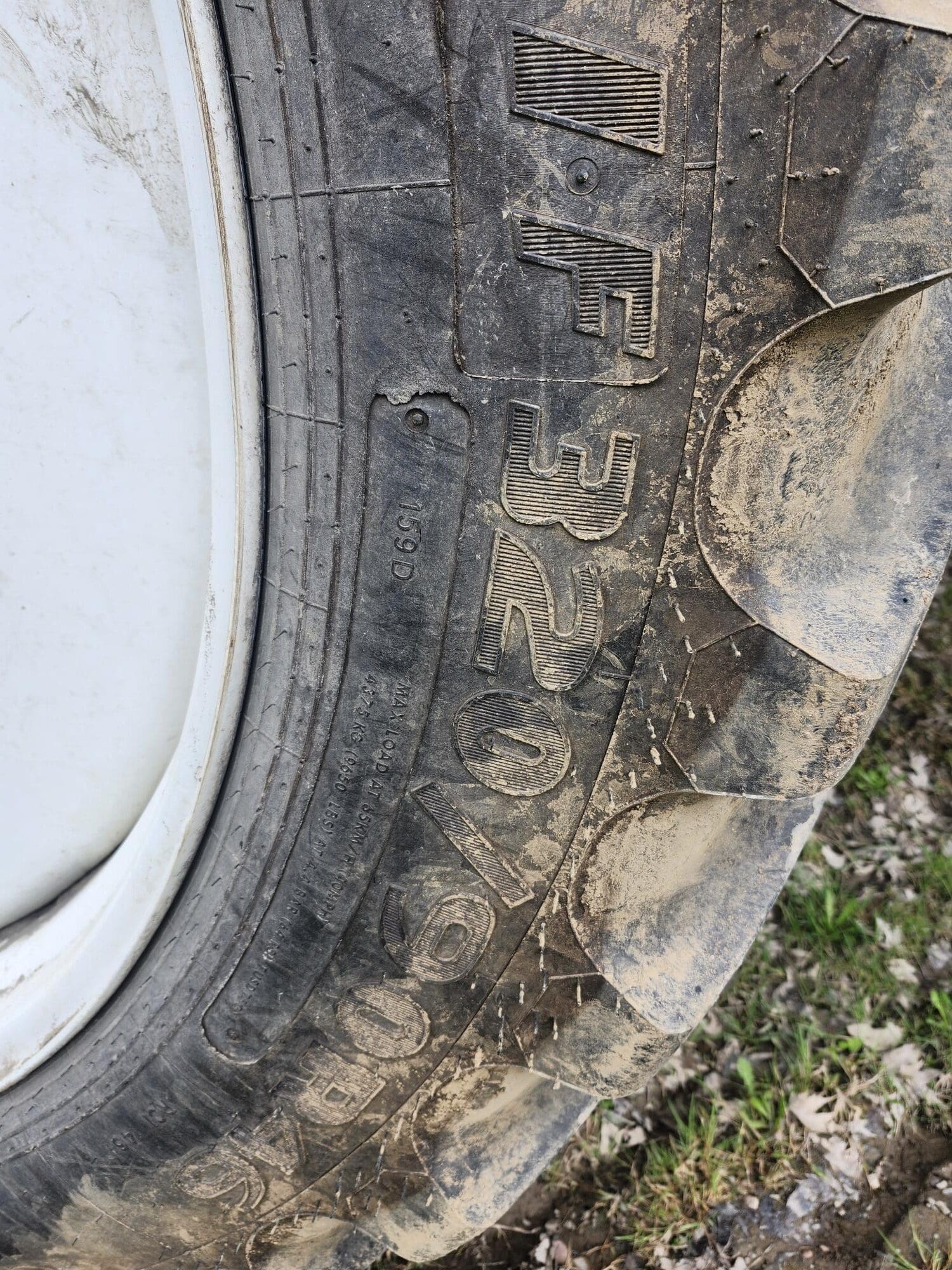 Gros plan d'un gros pneu de tracteur sale montrant la bande de roulement et les marquages en relief, notamment « IF 320/70R15 », sur un fond d'herbe et de terre.