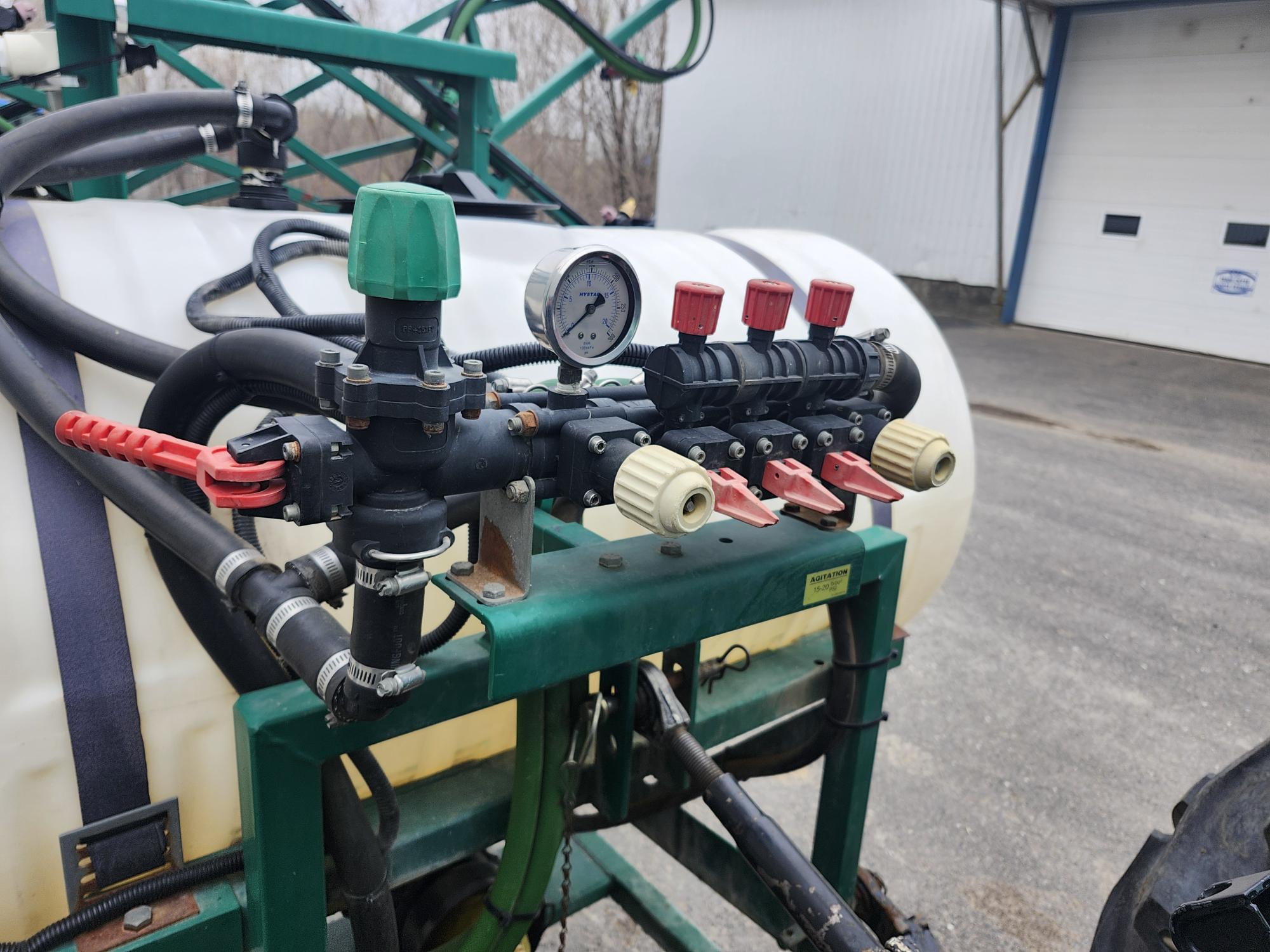 Gros plan d'un système de contrôle de pulvérisateur agricole avec manomètre, tuyaux et plusieurs poignées de vannes rouges, monté sur un cadre vert devant un grand réservoir blanc. Un bâtiment avec des portes de garage est visible en arrière-plan.