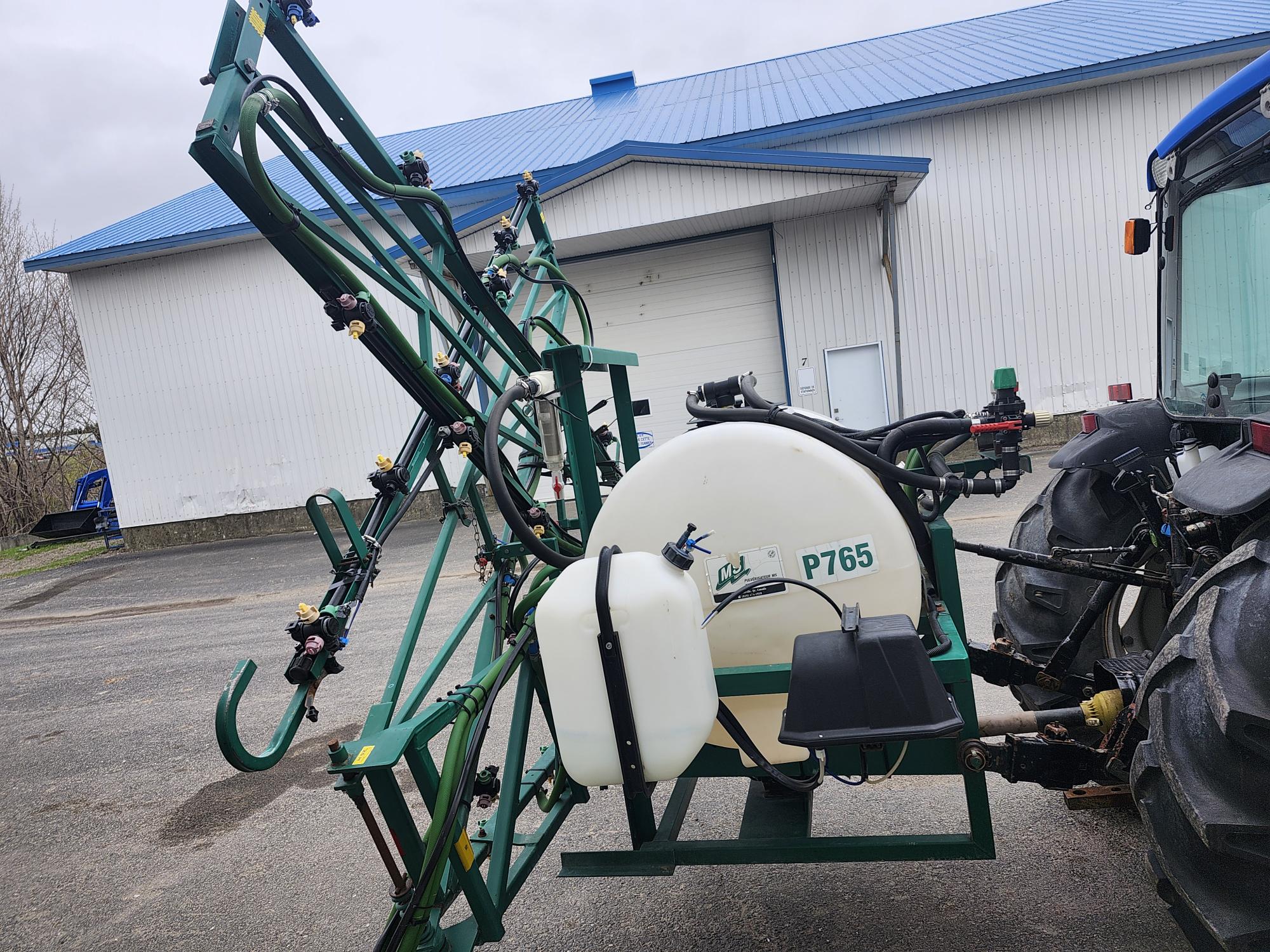 Un pulvérisateur agricole vert avec un réservoir blanc étiqueté « P765 » est attaché à l'arrière d'un tracteur, garé devant un grand bâtiment blanc avec un toit bleu.