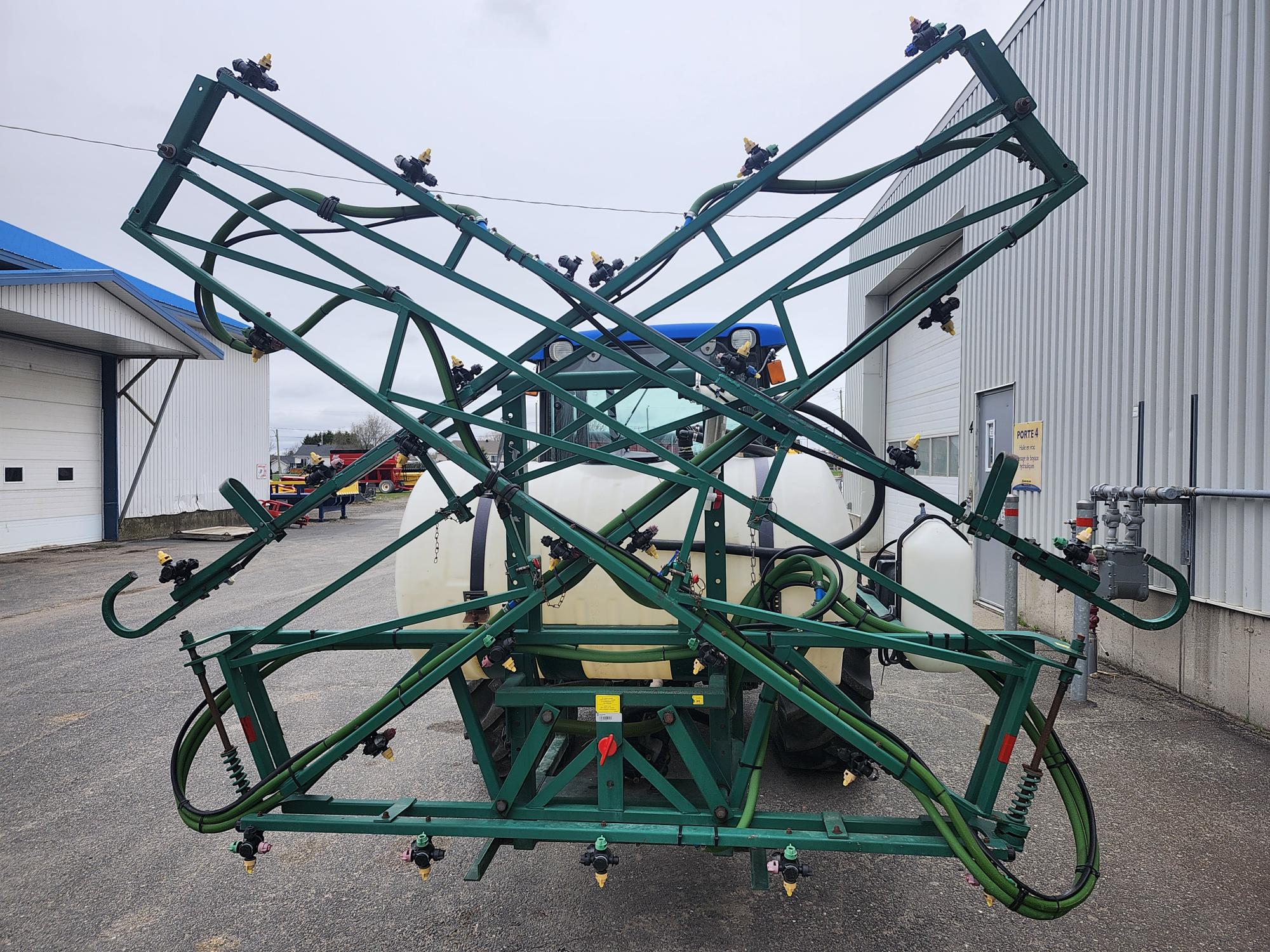 Un grand pulvérisateur agricole vert aux bras repliés est fixé à l'arrière d'un tracteur, garé sur un terrain en béton à côté de bâtiments industriels.