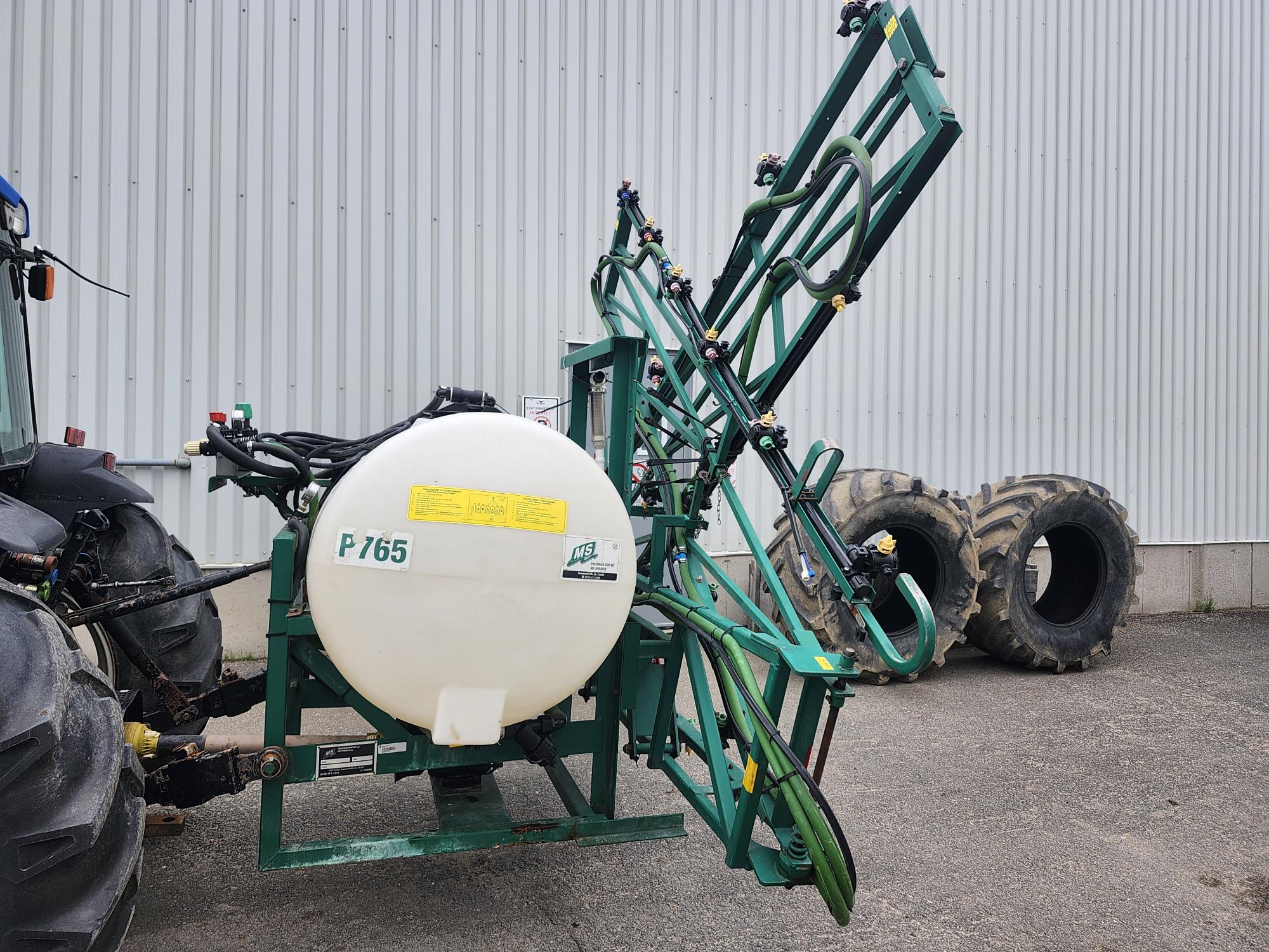 Un pulvérisateur agricole vert avec un réservoir blanc étiqueté « P 765 » est attelé à un tracteur. Deux gros pneus de tracteur sont empilés au sol à proximité, devant un bâtiment en tôle ondulée.
