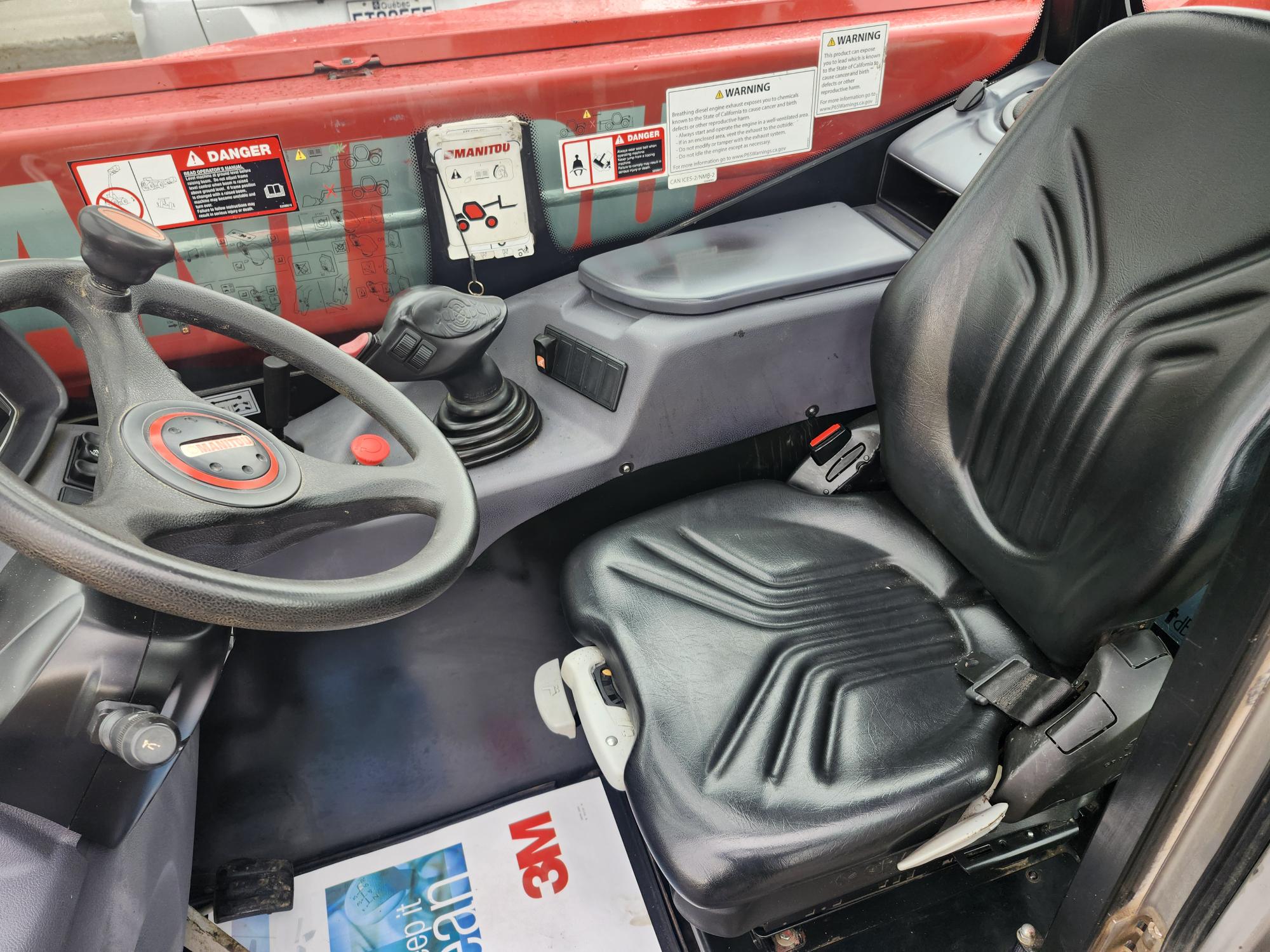 Gros plan de l'habitacle d'un véhicule, avec un siège conducteur noir, un volant, un levier de vitesse, des panneaux de commande avec des étiquettes d'avertissement et un magazine 3M posé au sol. Le véhicule semble être un véhicule industriel ou de construction.
