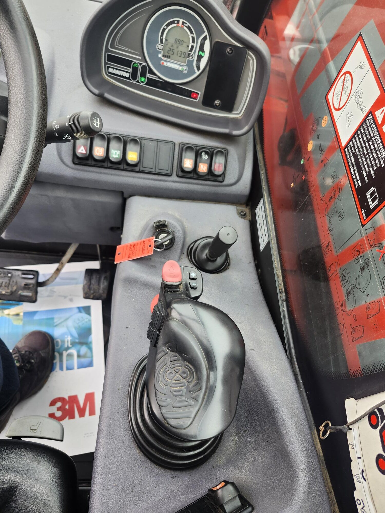 Tableau de commande d'un véhicule ou d'une machine lourde avec boutons, interrupteurs, joystick, compteur de vitesse et étiquettes d'avertissement. On y voit également une partie du volant, une clé et le pied d'une personne.