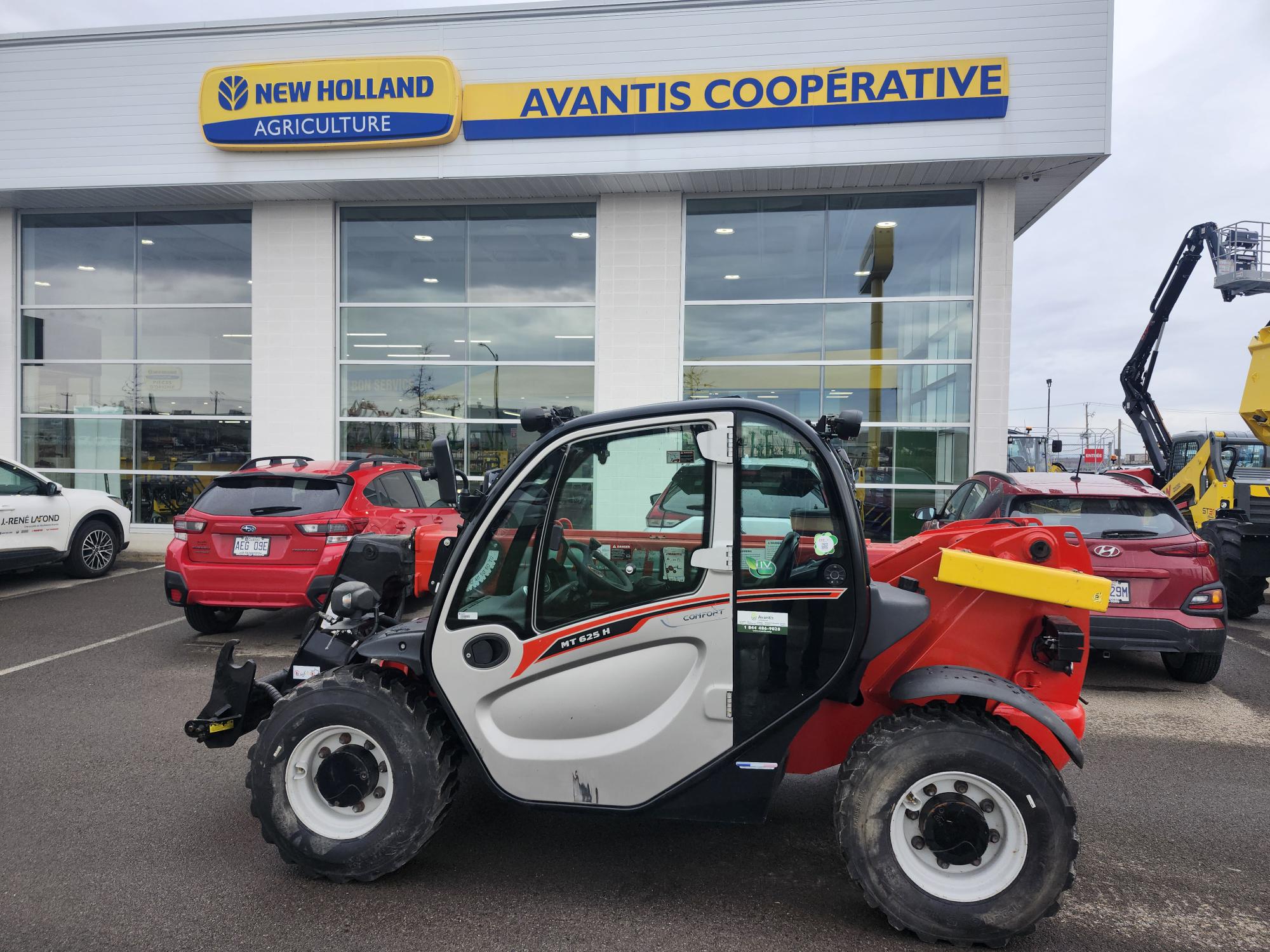 Un chariot télescopique compact rouge et blanc est stationné à l'extérieur d'un concessionnaire Avantis Coopérative New Holland Agriculture, avec plusieurs voitures rouges et de grandes fenêtres en arrière-plan.