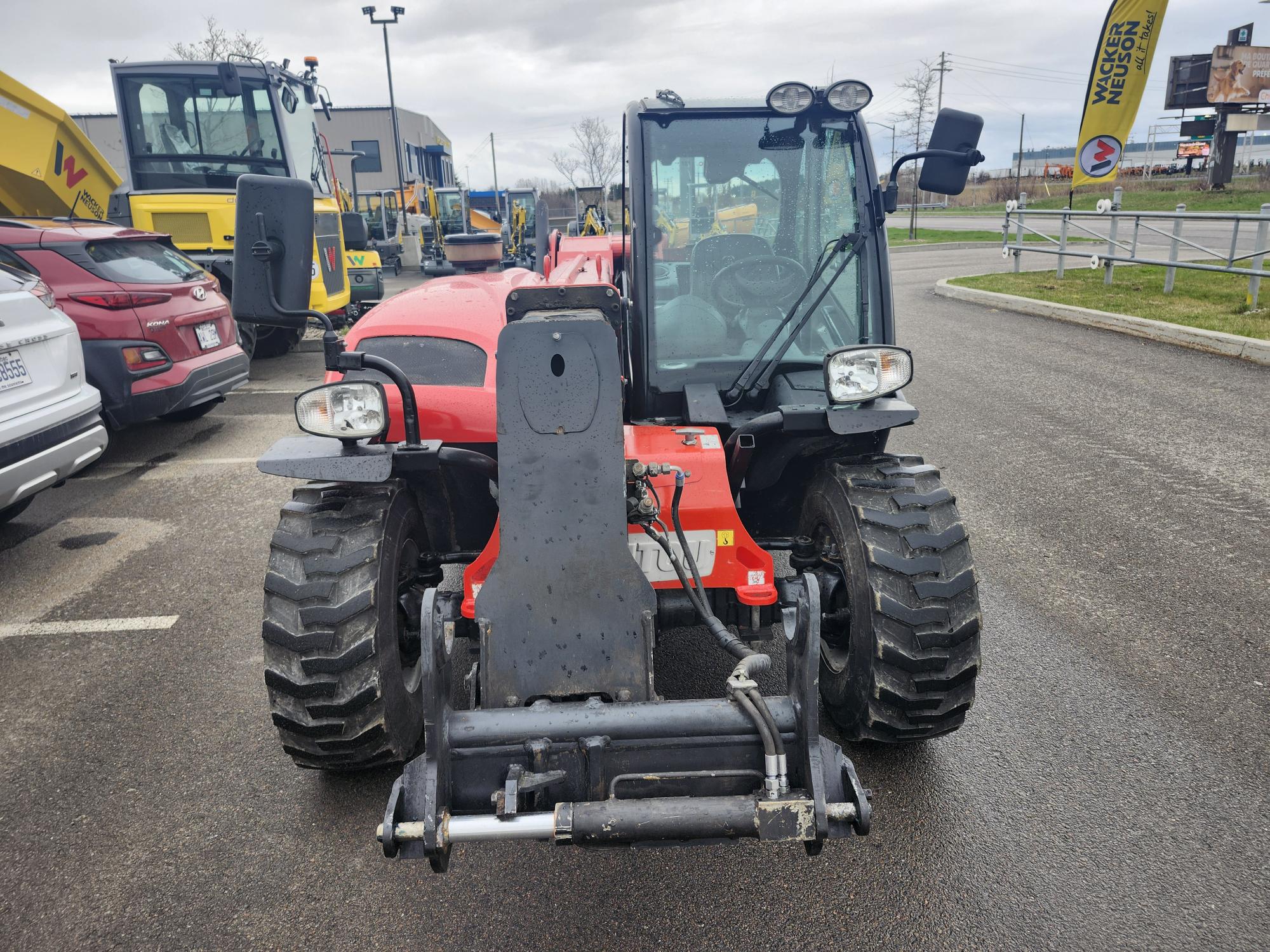 Un chariot télescopique rouge, également connu sous le nom de chariot télescopique, est garé sur un terrain entouré d'autres véhicules et d'équipements de construction par une journée nuageuse.