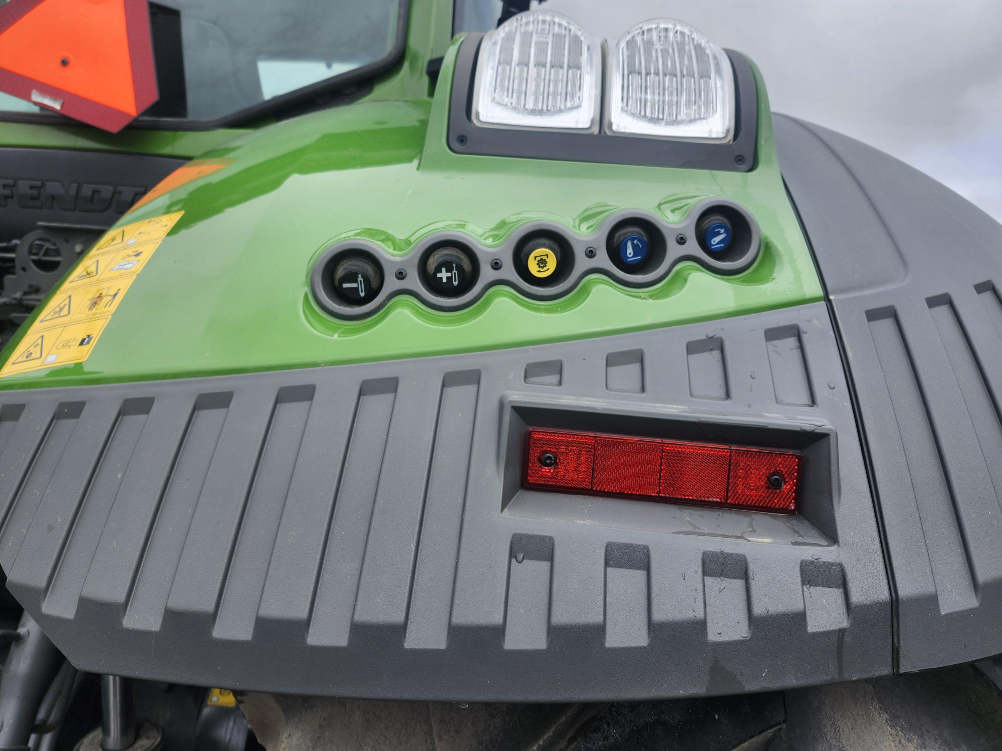 Gros plan d'un véhicule vert et gris, peut-être un tracteur, montrant des boutons de commande, des étiquettes d'avertissement, un réflecteur rouge et deux grands phares sur le dessus contre un ciel nuageux.