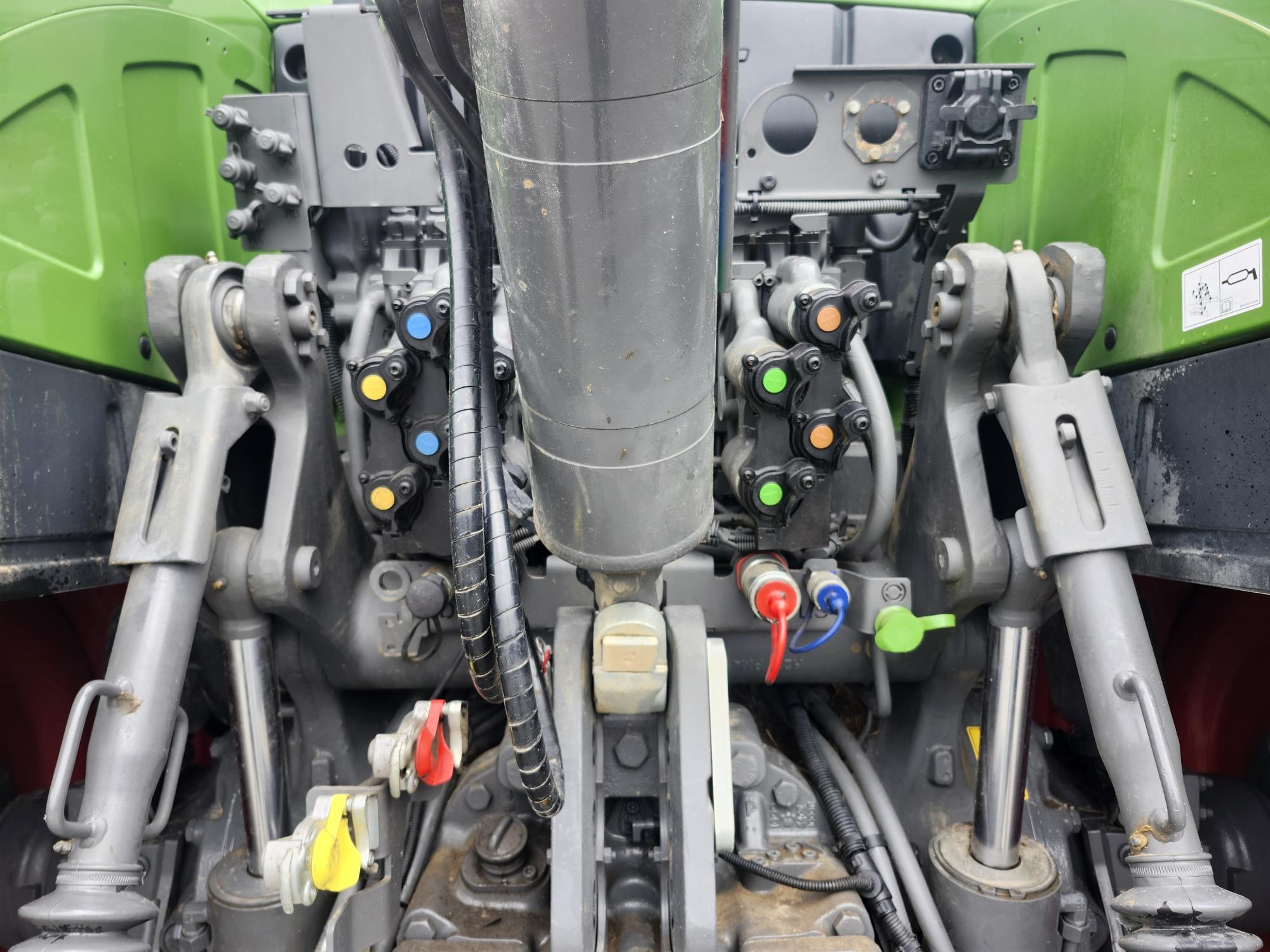 Vue rapprochée des connexions et des commandes hydrauliques arrière d'un tracteur vert, montrant des bras métalliques, des coupleurs hydrauliques colorés, des tuyaux et des leviers utilisés pour attacher et faire fonctionner les outils agricoles.