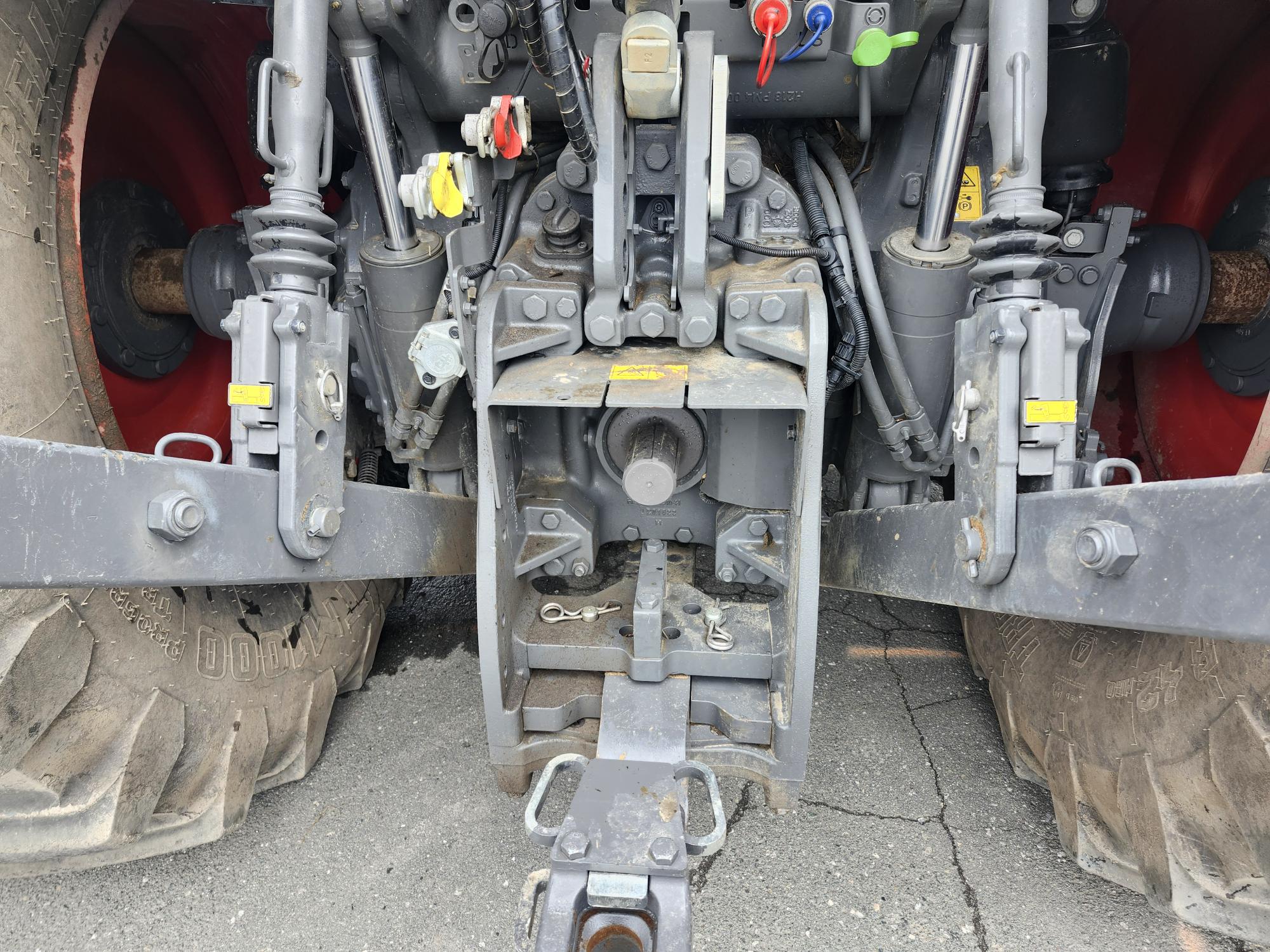 Gros plan de l'attelage arrière et des connexions hydrauliques d'un tracteur, montrant des pièces mécaniques, des leviers, des capuchons de commande colorés, de gros pneus arrière et une section de chaussée fissurée en dessous.