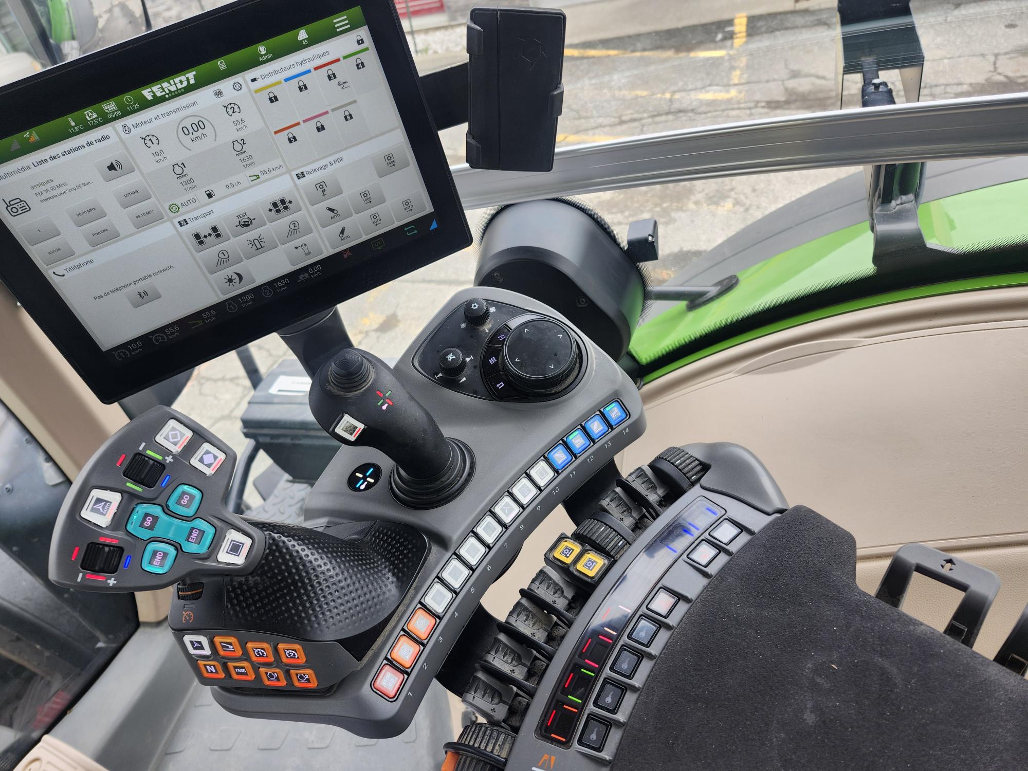 Vue rapprochée d'un cockpit de tracteur moderne, montrant un écran tactile avec des commandes, plusieurs boutons, des leviers et un joystick sur un tableau de bord, avec une partie d'un siège beige visible.