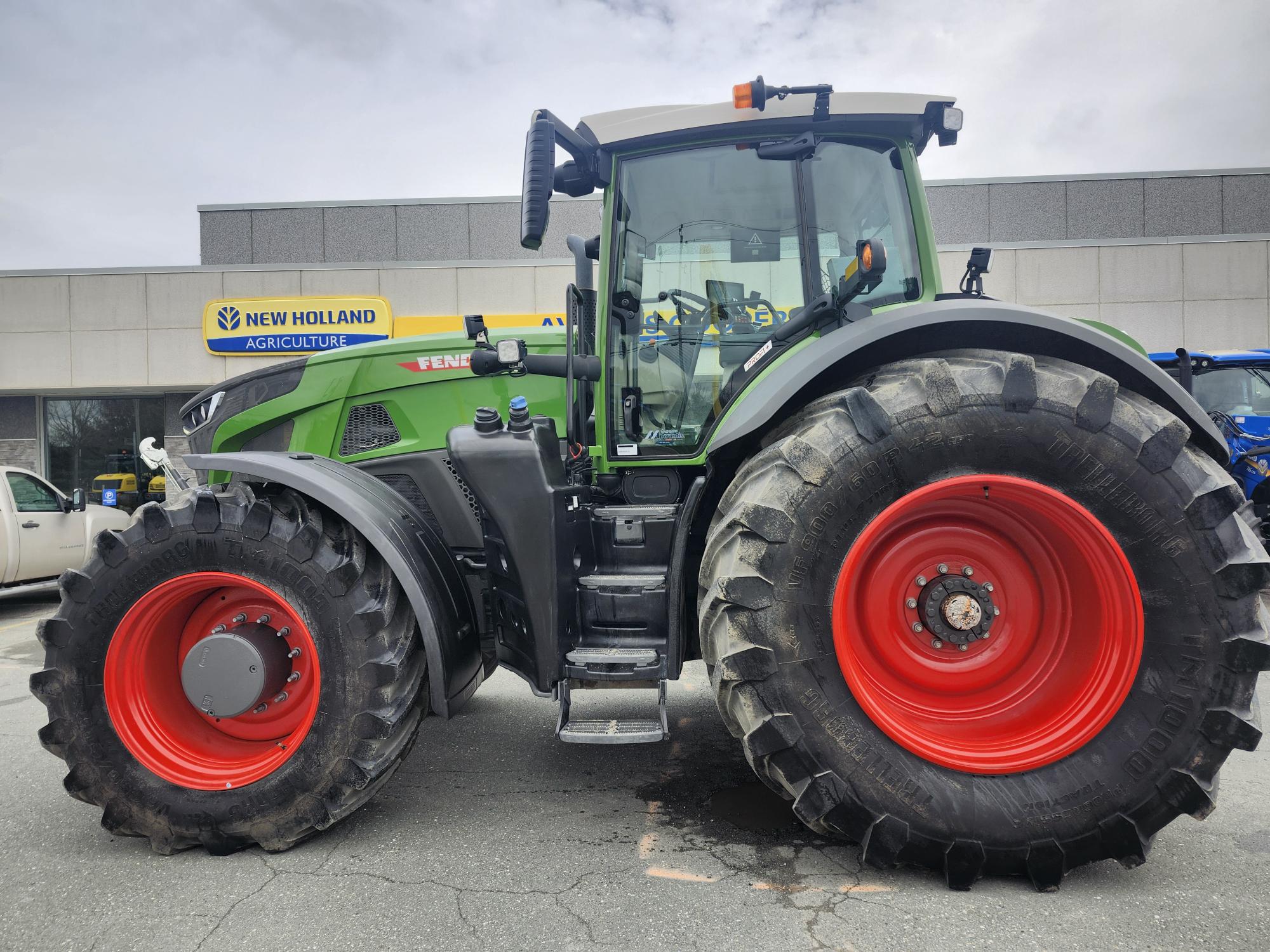 Un grand tracteur Fendt vert avec des roues rouges est garé à l'extérieur d'un bâtiment de concession New Holland Agriculture par une journée nuageuse.