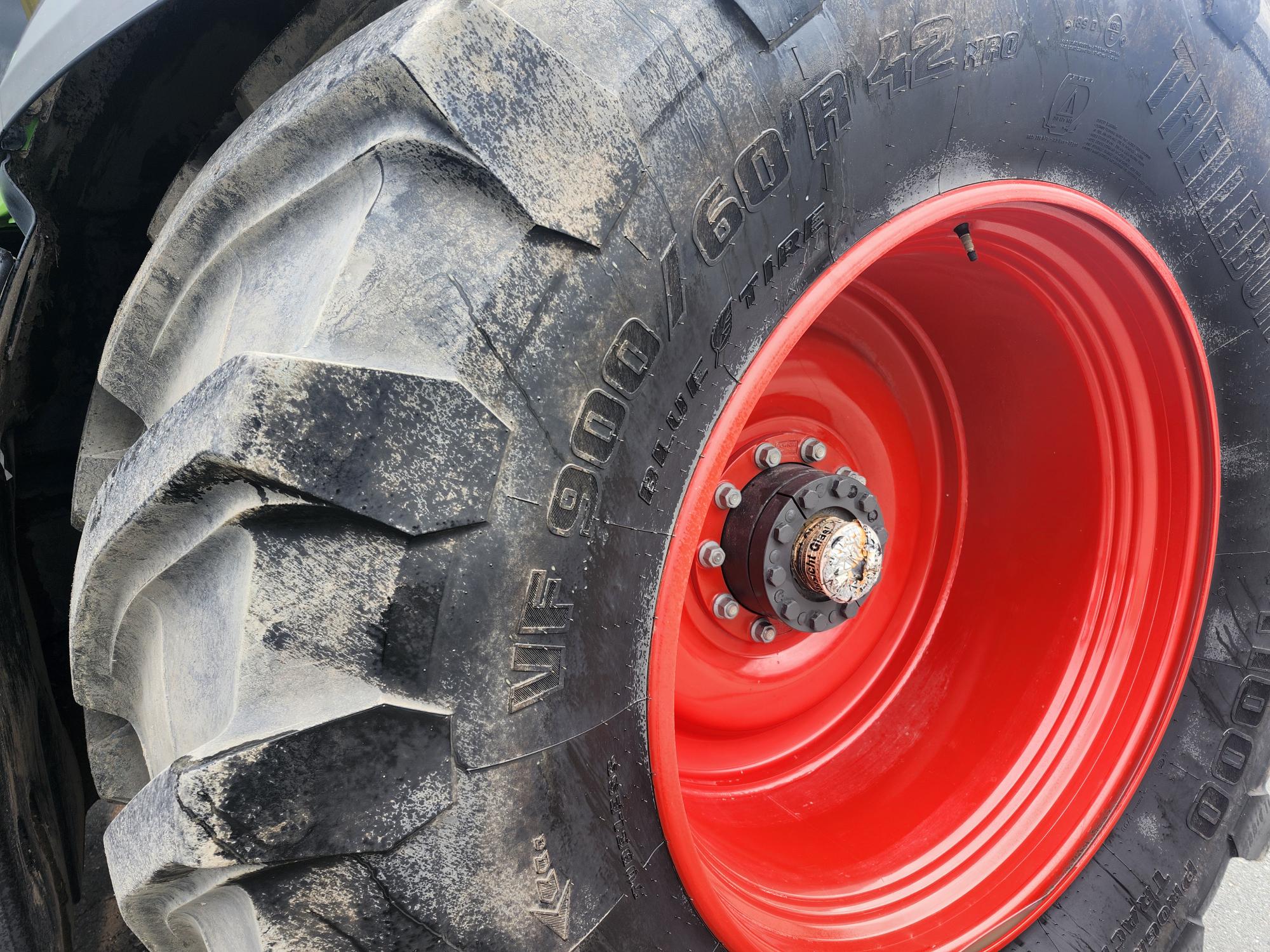 Gros plan sur un gros pneu de tracteur avec une jante rouge. La bande de roulement est usée et poussiéreuse, et des boulons sont visibles au centre de la roue. La taille et la marque du pneu sont clairement indiquées sur le flanc.
