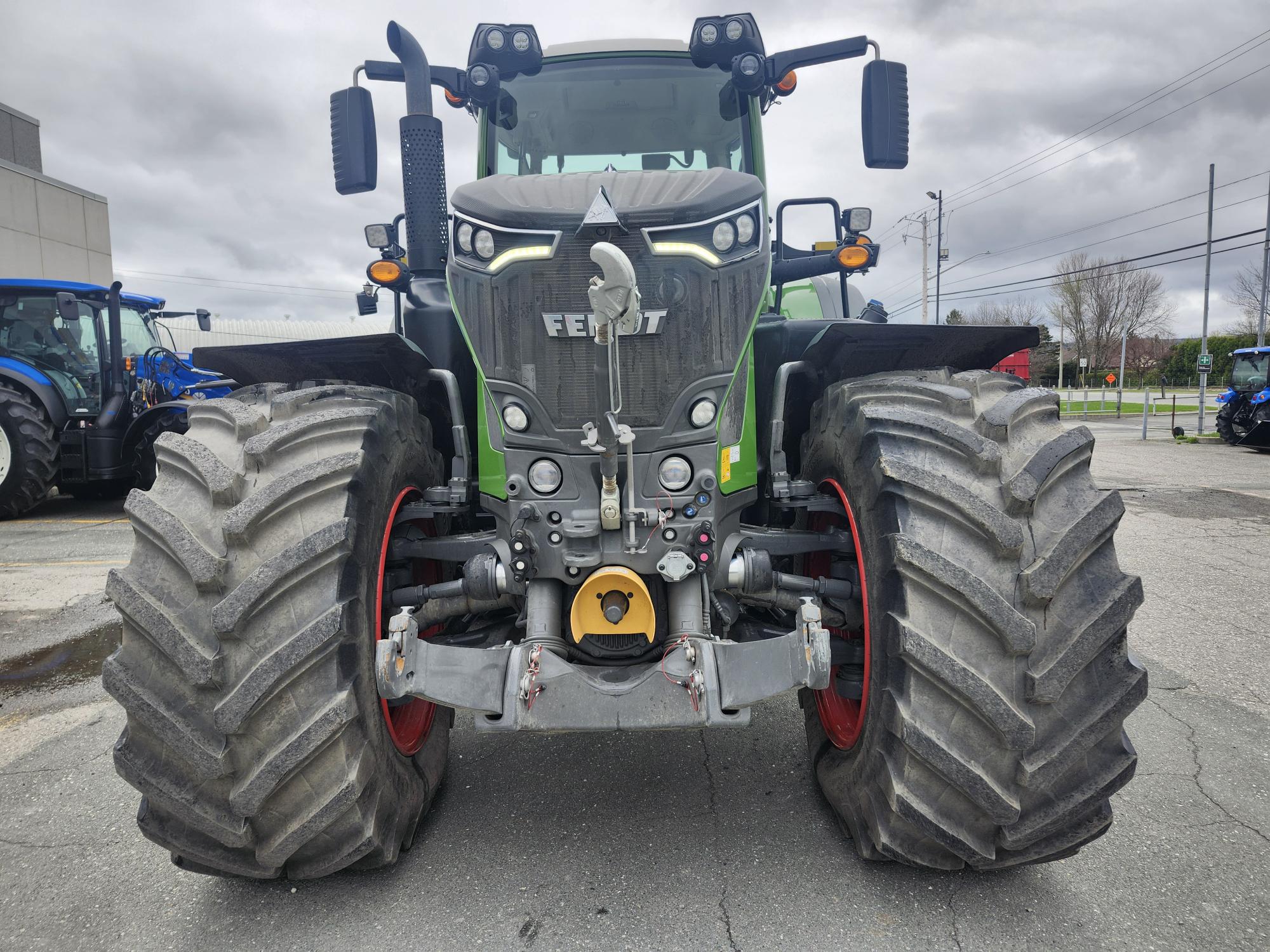 Vue de face d'un grand tracteur Fendt vert avec des pneus doubles des deux côtés, stationné sur une surface pavée avec un ciel nuageux et d'autres machines visibles en arrière-plan.