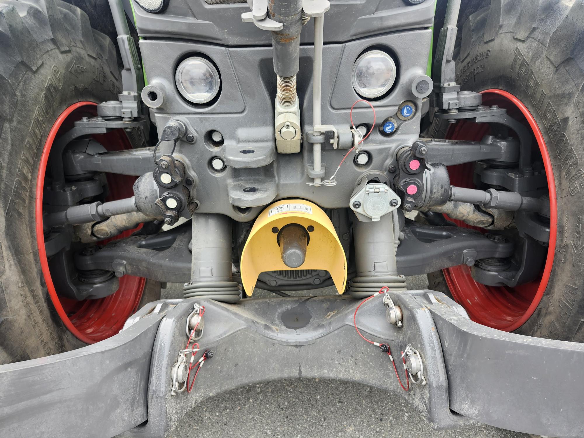 Gros plan de l'arrière d'un tracteur montrant les composants mécaniques et hydrauliques, le câblage et les points de connexion. De grands pneus noirs à jantes rouges sont visibles de chaque côté.