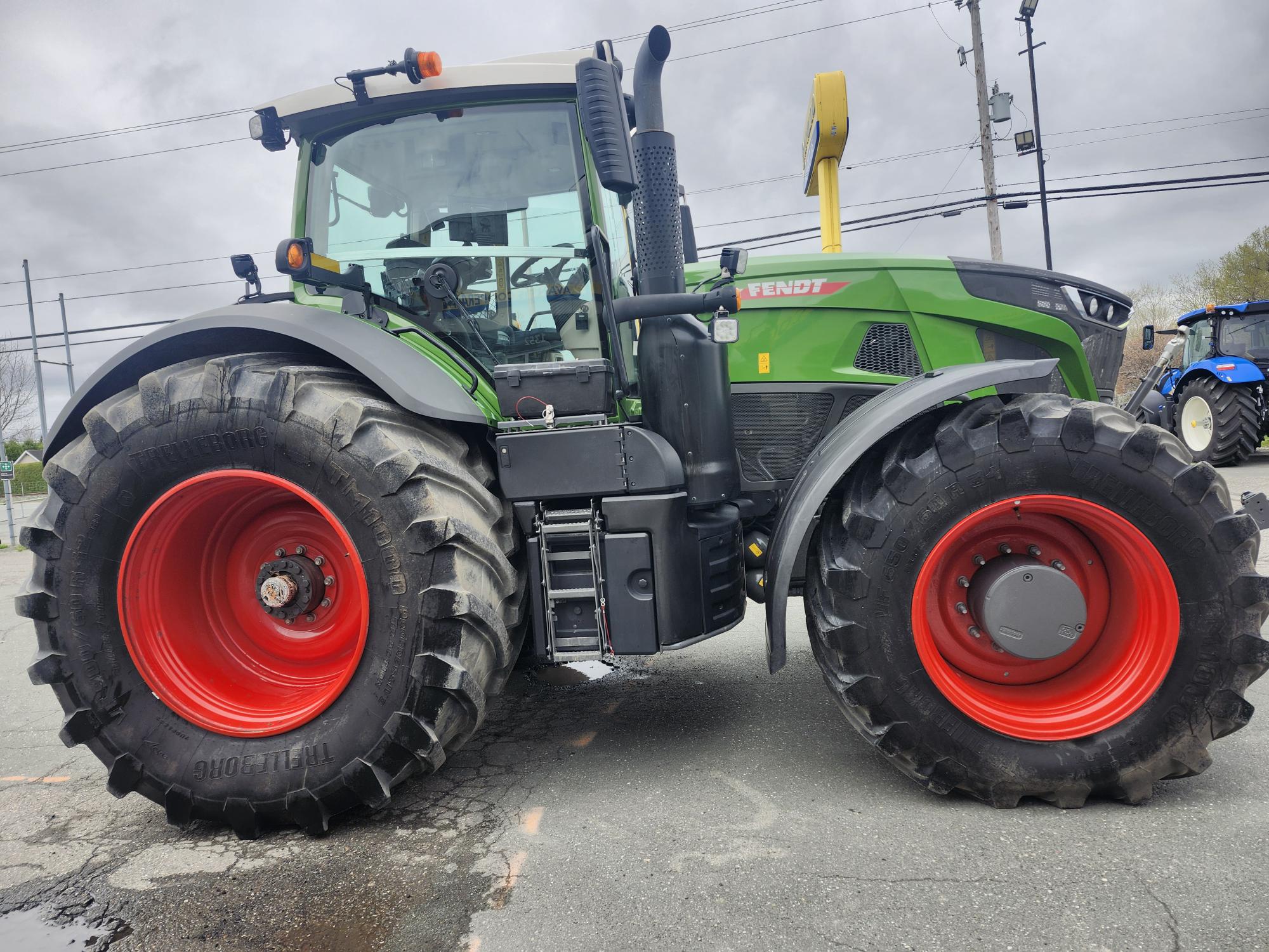 Un gros tracteur Fendt vert aux pneus noirs et rouges est garé sur un terrain pavé sous un ciel nuageux. Des lignes électriques et un autre tracteur bleu sont visibles en arrière-plan.