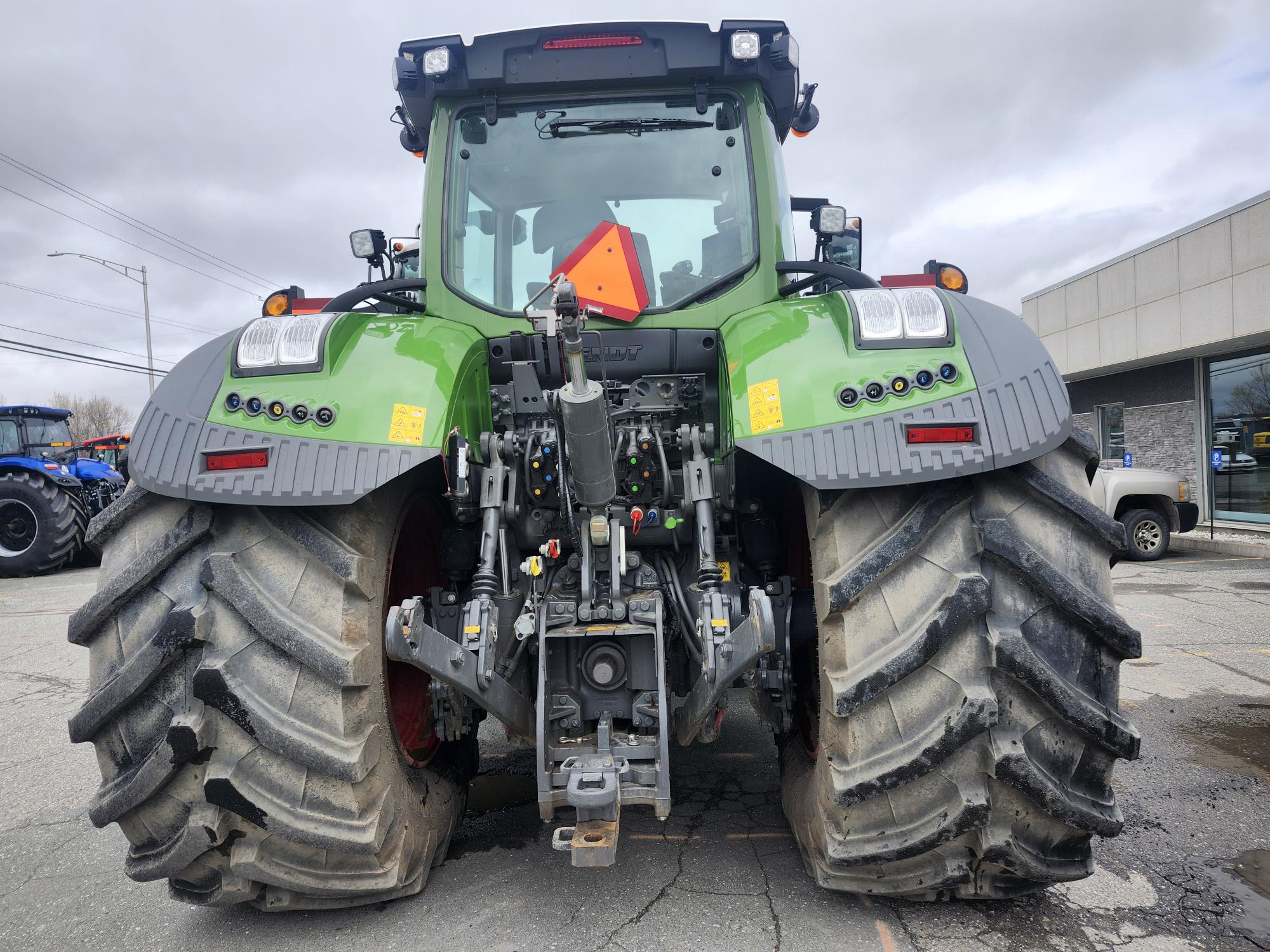 Un gros tracteur vert vu de l'arrière sur un terrain pavé, montrant ses larges pneus boueux, son attelage arrière et son triangle de présignalisation. Un ciel couvert et d'autres véhicules sont visibles en arrière-plan.