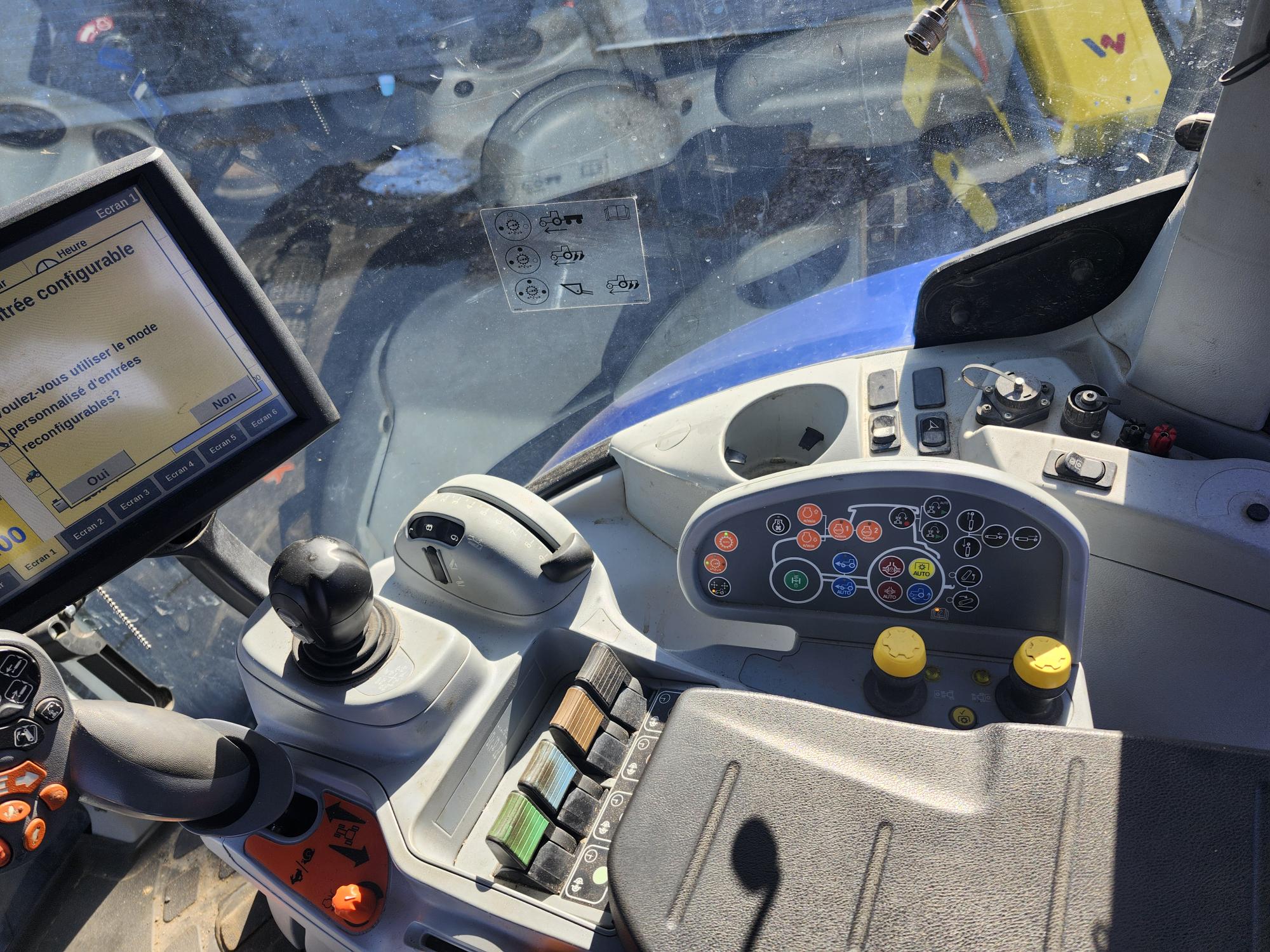 Gros plan de l'intérieur de la cabine d'un tracteur montrant un affichage numérique, un panneau de commande avec divers boutons, joysticks et interrupteurs, et une vue d'un autre véhicule à travers le pare-brise avant.