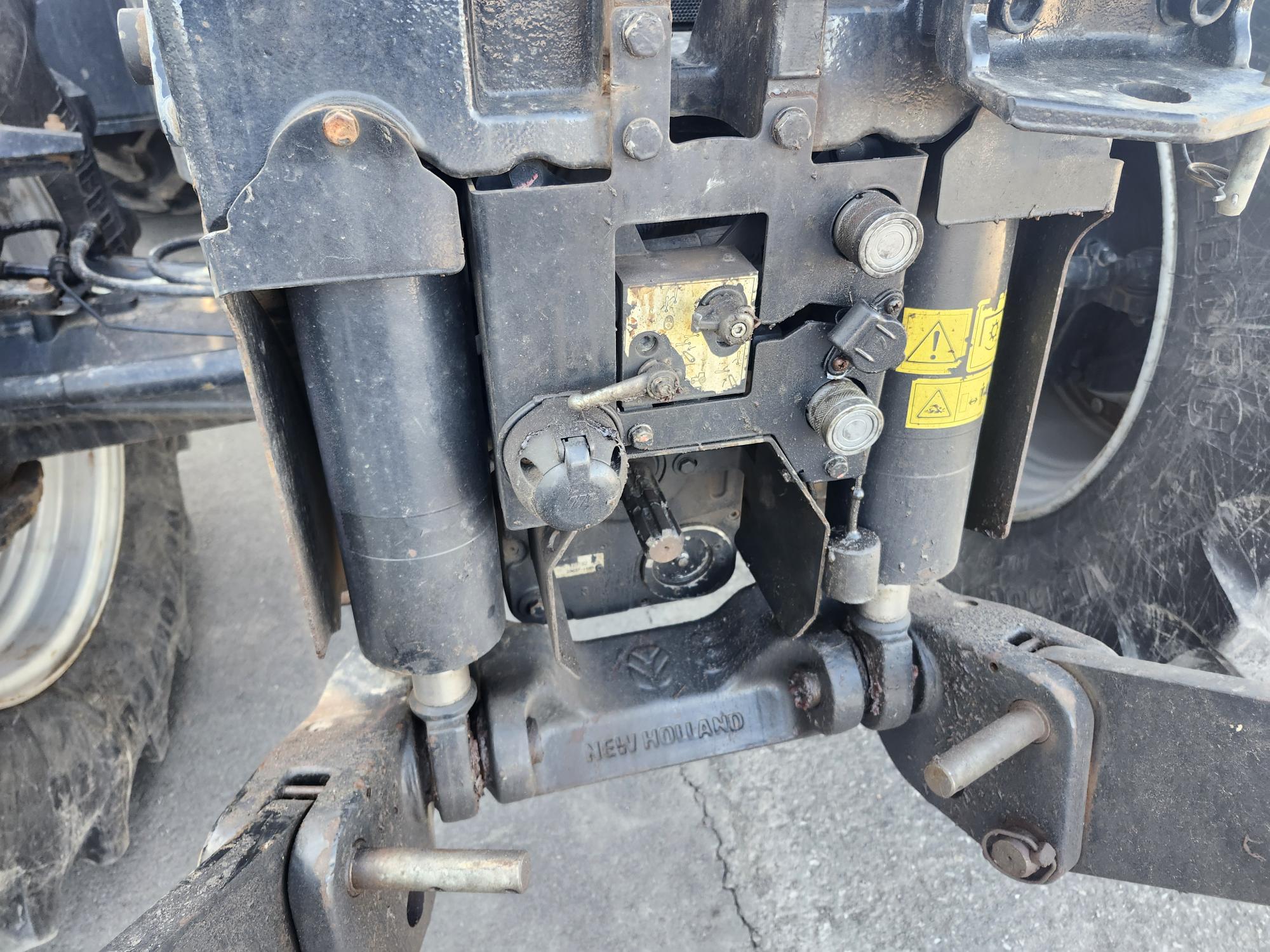 Gros plan d'une pièce mécanique sur un tracteur New Holland, montrant divers leviers, boulons, composants hydrauliques, autocollants d'avertissement et une partie de la roue arrière en arrière-plan.