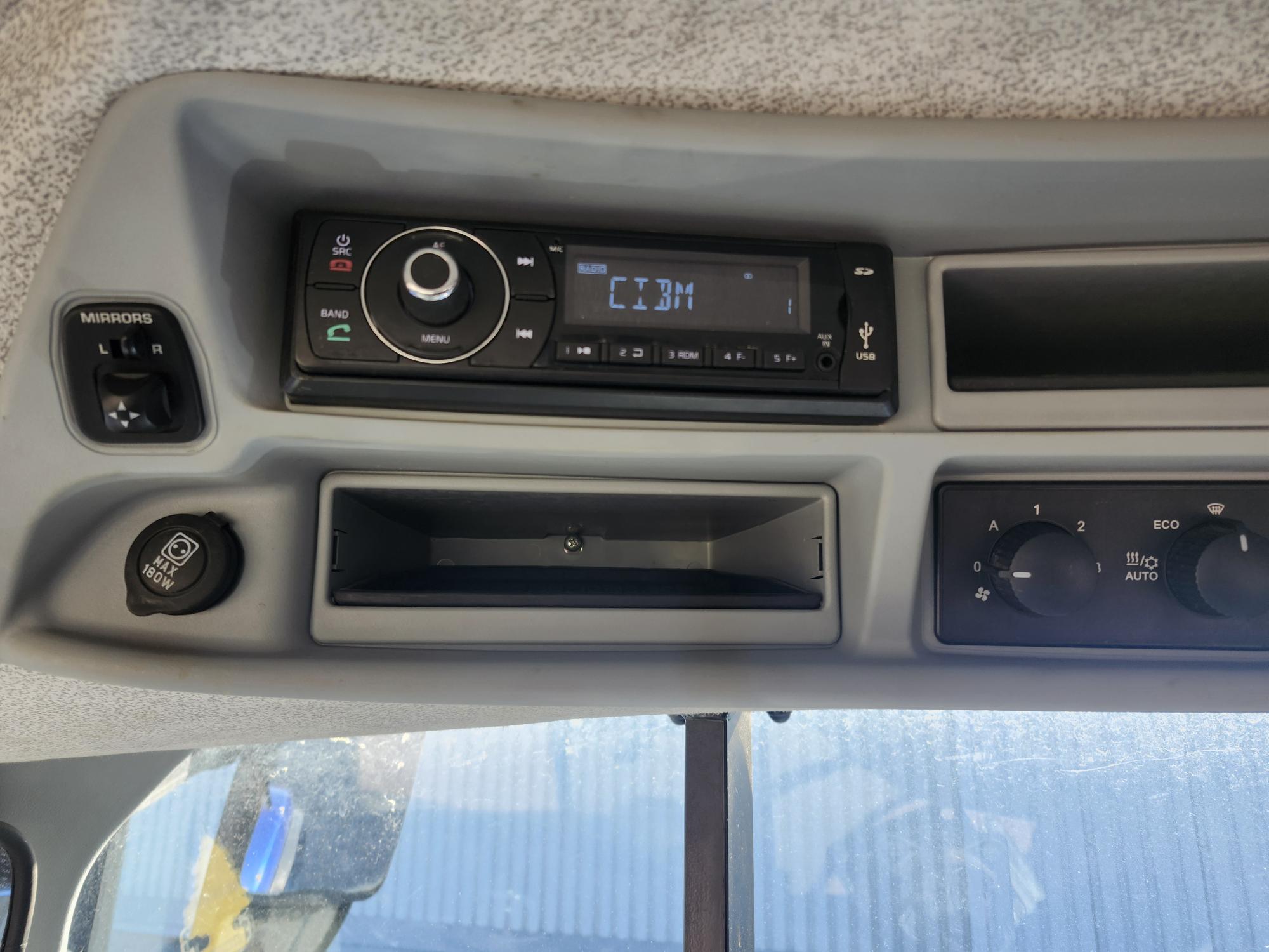 Tableau de bord d'un véhicule montrant un écran radio indiquant « 12:30 », des boutons de contrôle de la climatisation, un petit compartiment de rangement, un interrupteur de réglage des rétroviseurs et une partie du pare-brise avec la lumière du soleil visible.