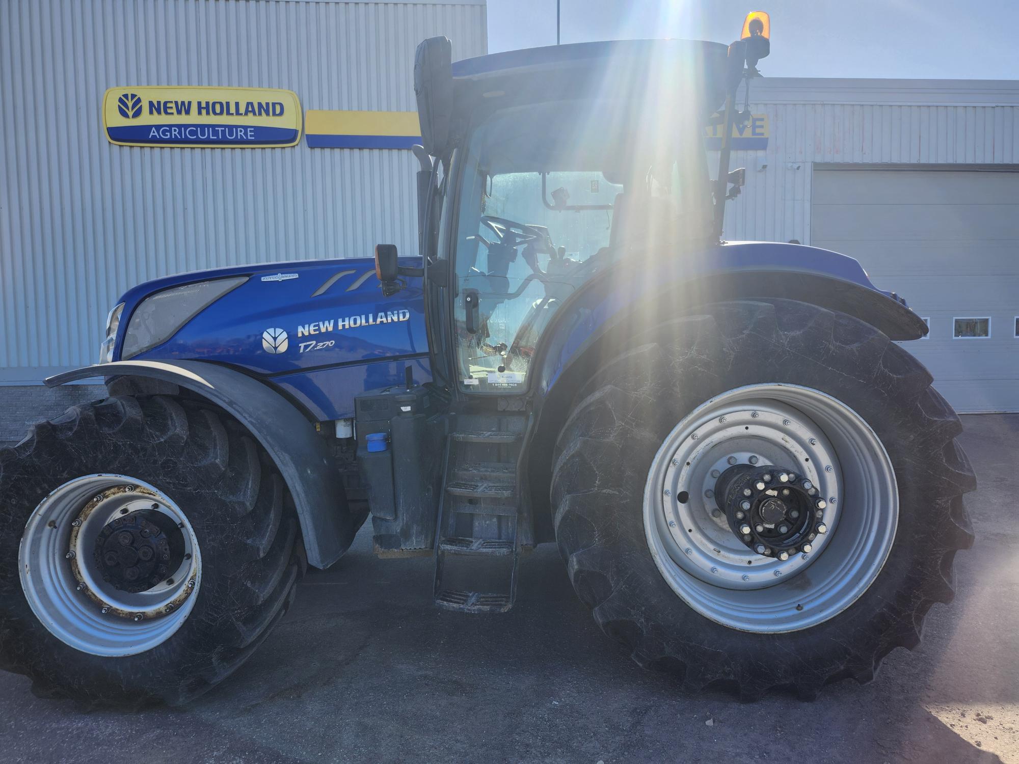 Un tracteur bleu New Holland T7.210 est garé à l'extérieur d'un bâtiment agricole New Holland, la lumière du soleil créant un éblouissement sur l'image.