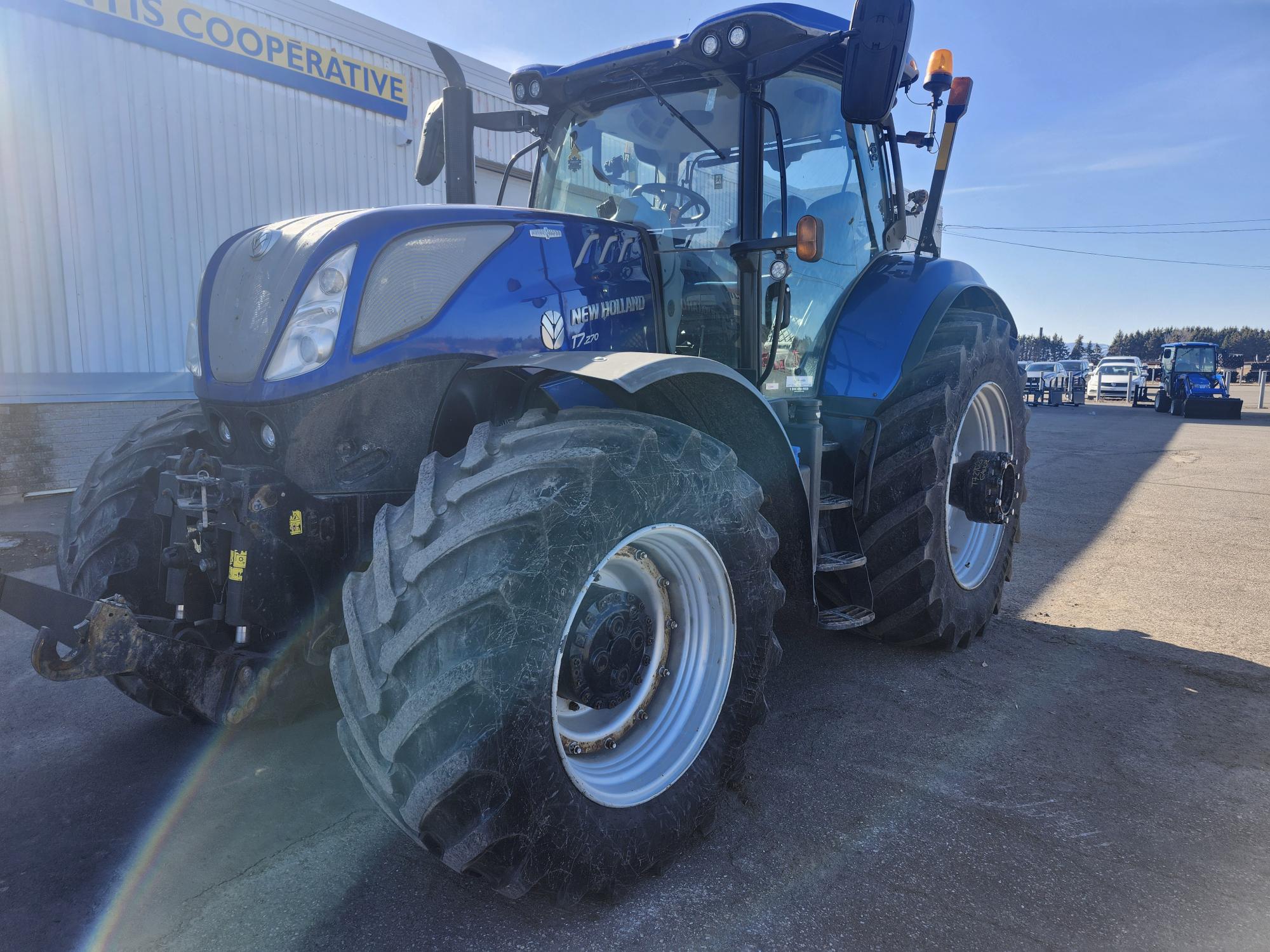 Un gros tracteur New Holland bleu aux pneus surdimensionnés est garé sur un terrain pavé, baigné de soleil, près d'un bâtiment portant l'inscription « COOPÉRATIVE ». D'autres véhicules sont visibles en arrière-plan.