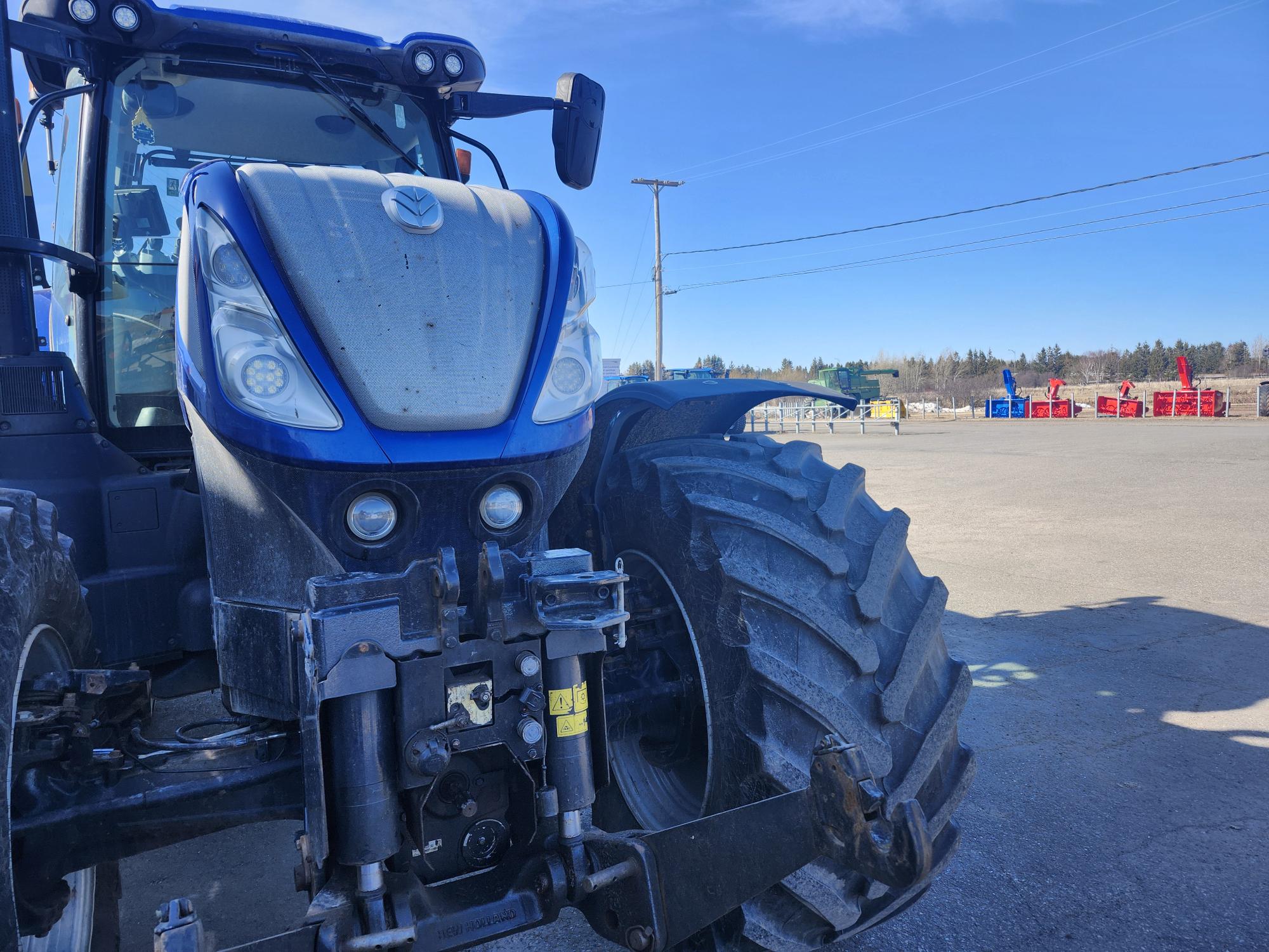 Vue rapprochée d'un gros tracteur bleu stationné sur un terrain asphalté, avec des équipements industriels et des conteneurs rouges visibles en arrière-plan sous un ciel bleu clair.