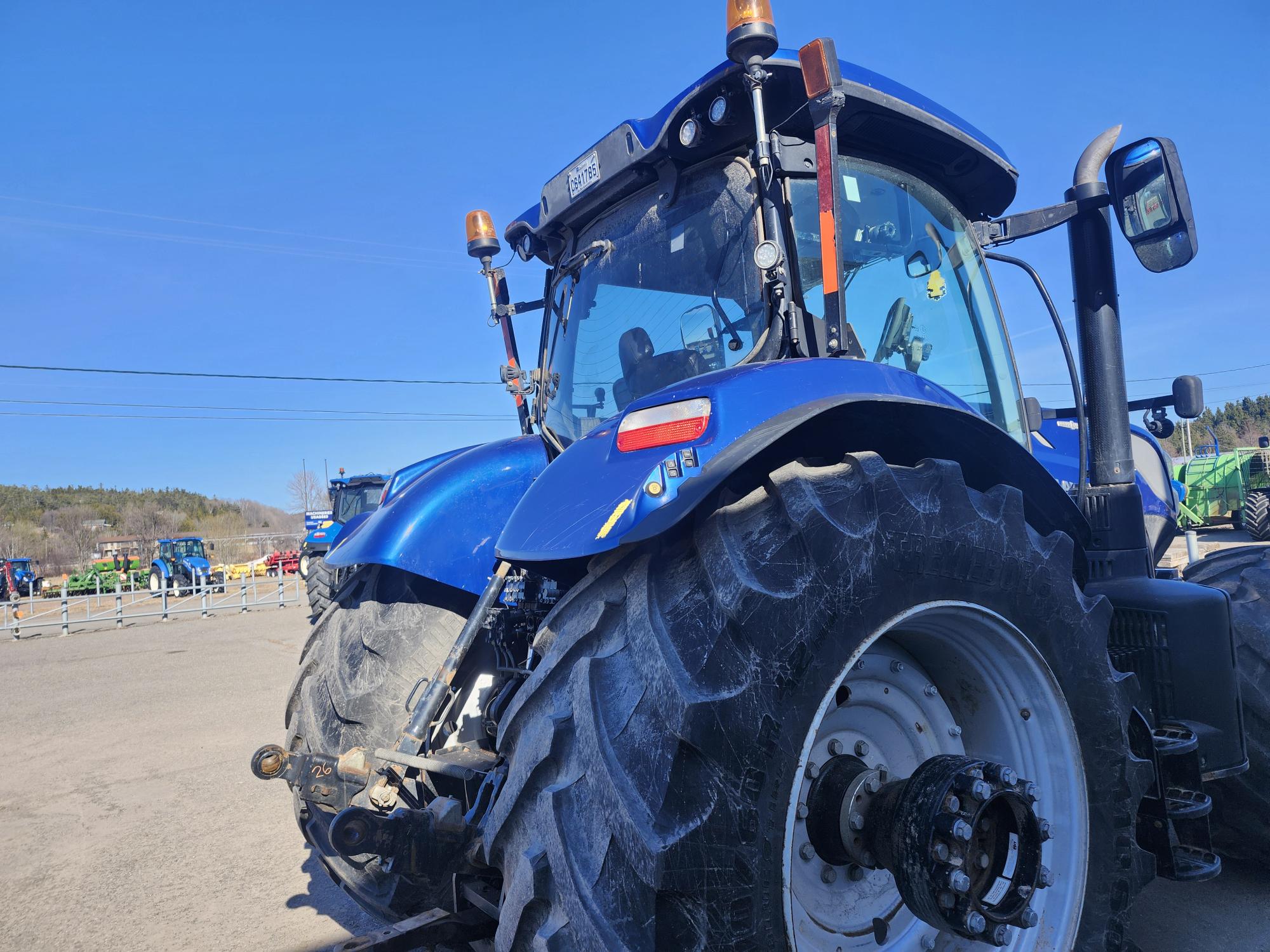 Gros plan d'un tracteur bleu garé sur un terrain pavé, avec de gros pneus arrière et un ciel bleu clair au-dessus. D'autres équipements agricoles et des clôtures sont visibles en arrière-plan.