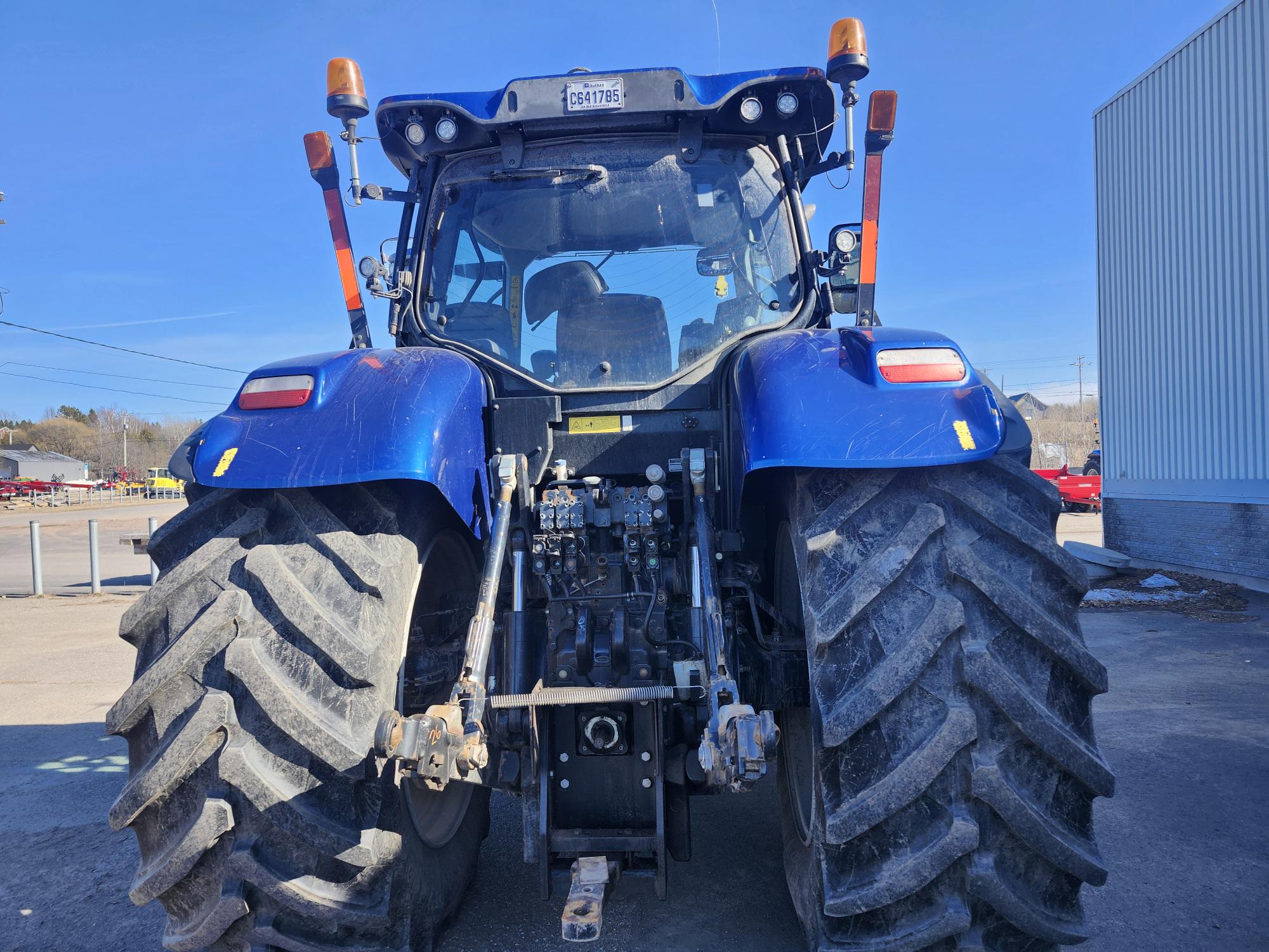 Un tracteur bleu est garé dehors par une journée ensoleillée. Vu de l'arrière, il laisse apparaître de gros pneus sales et des composants mécaniques. Des bâtiments et des véhicules sont visibles en arrière-plan.