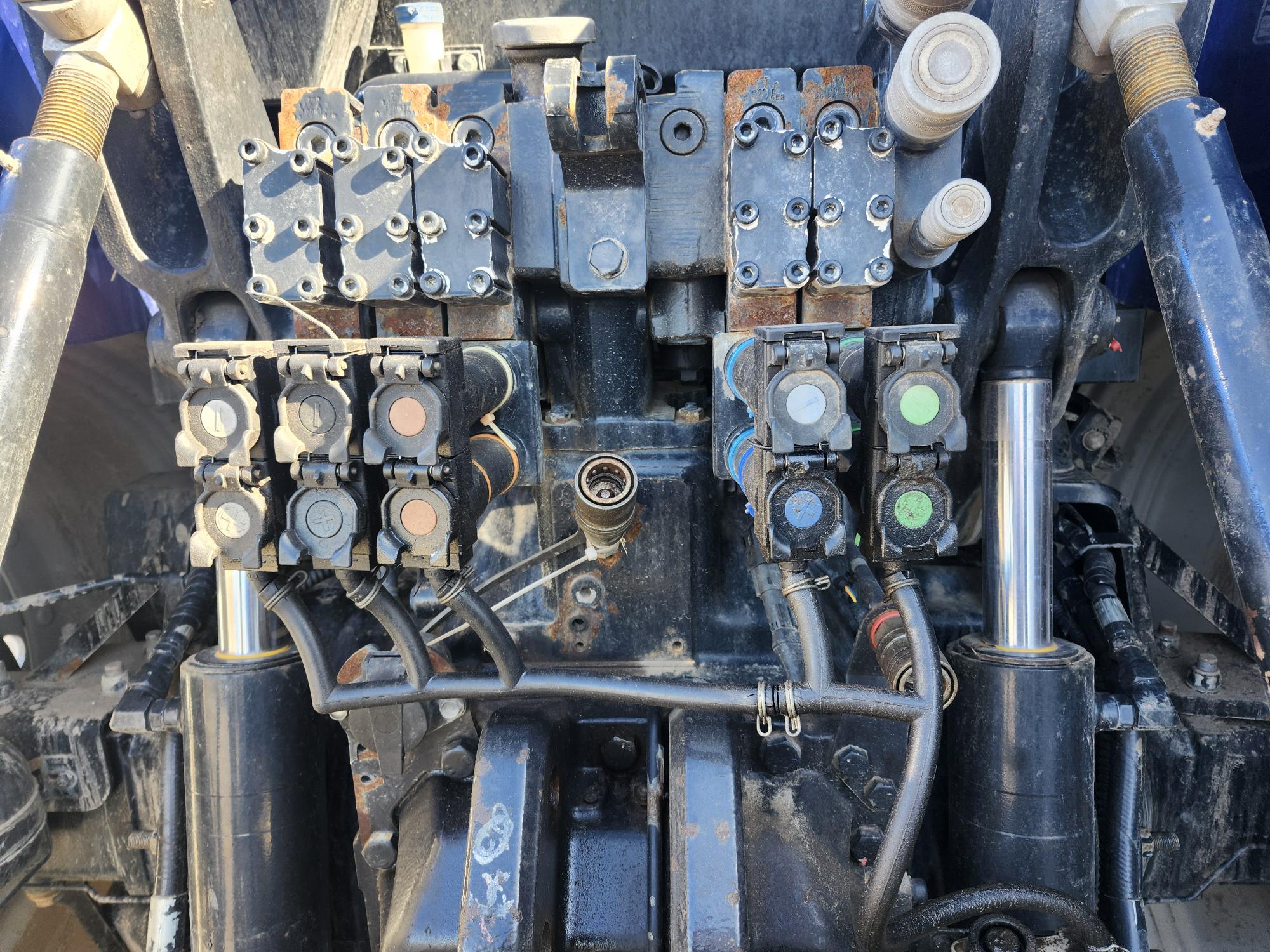 Vue rapprochée des connecteurs et raccords hydrauliques sur le panneau arrière d'un tracteur, montrant plusieurs ports et tuyaux, avec de la poussière et de la rouille visibles sur les pièces métalliques.