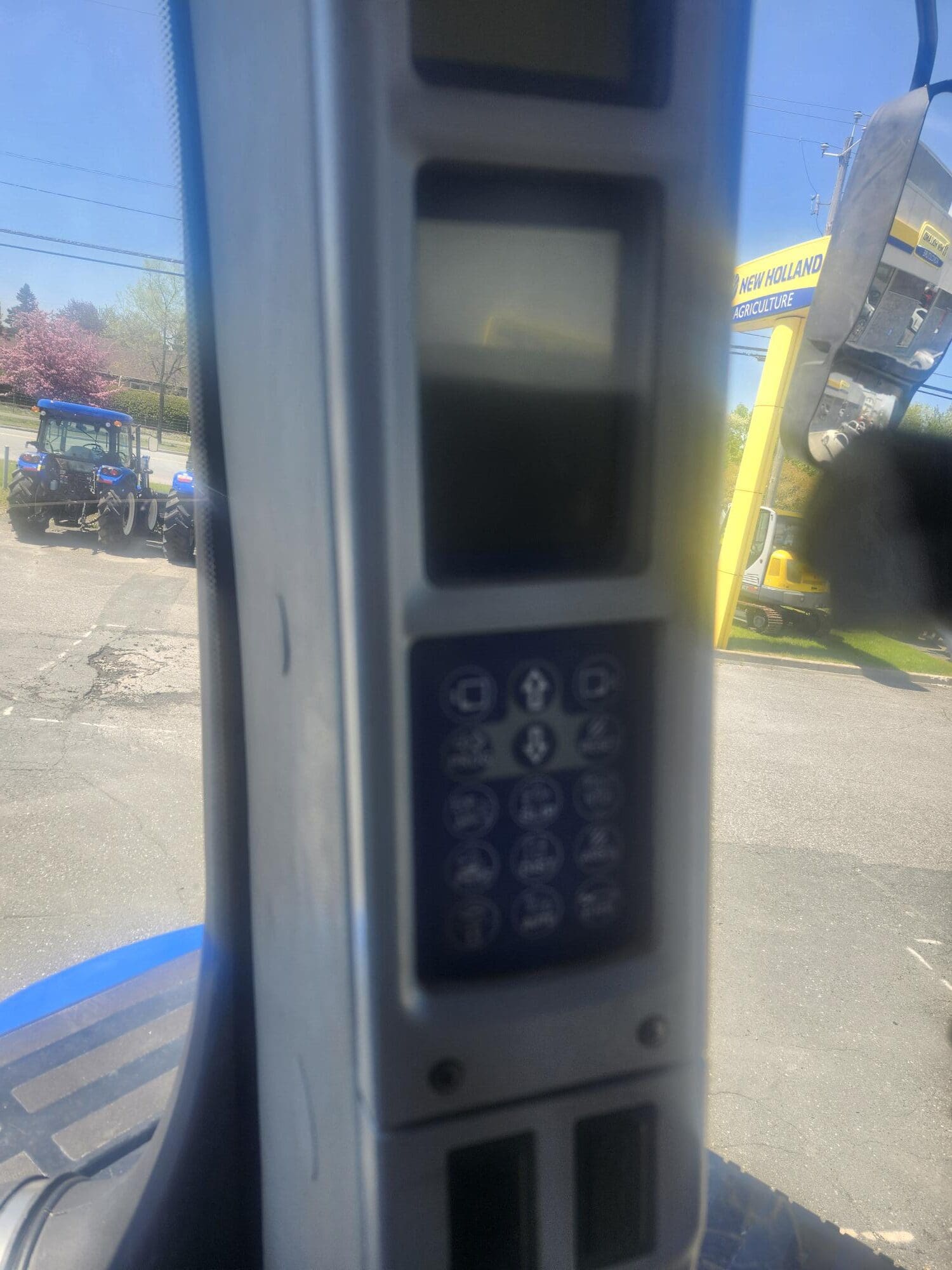 Gros plan d'un panneau de commande avec boutons et écran d'affichage, situé à l'intérieur d'un véhicule. À l'extérieur, un tracteur et un panneau jaune New Holland Agriculture sont visibles sous un ciel dégagé.