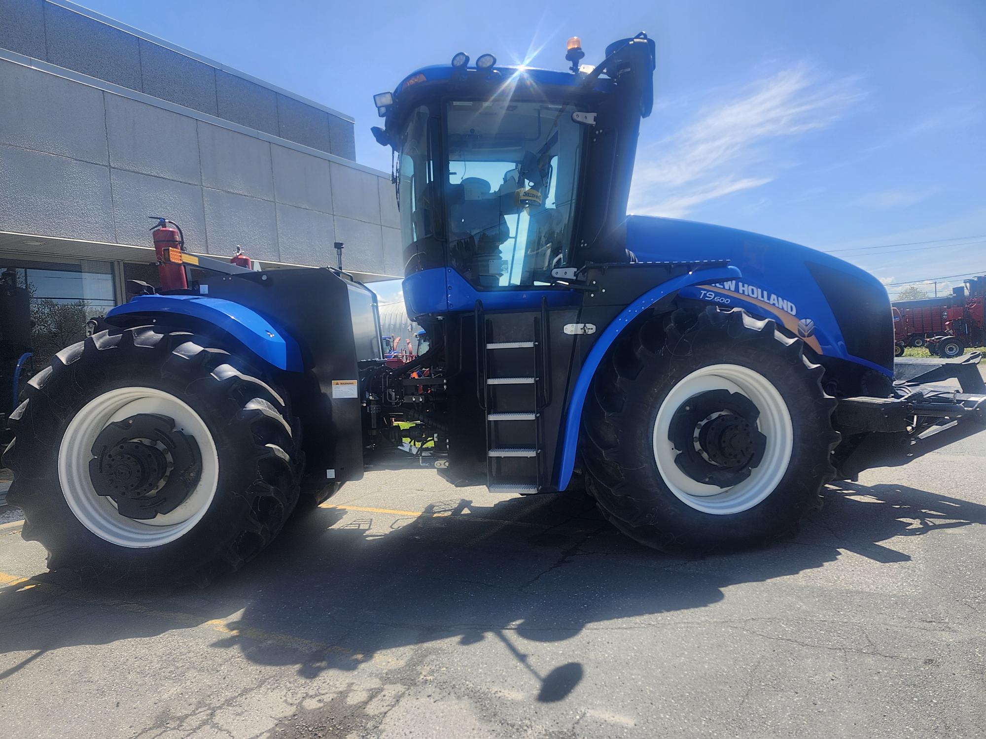 Un gros tracteur bleu New Holland est garé à l'extérieur par une journée ensoleillée devant un bâtiment commercial, avec ses gros pneus et sa cabine clairement visibles.