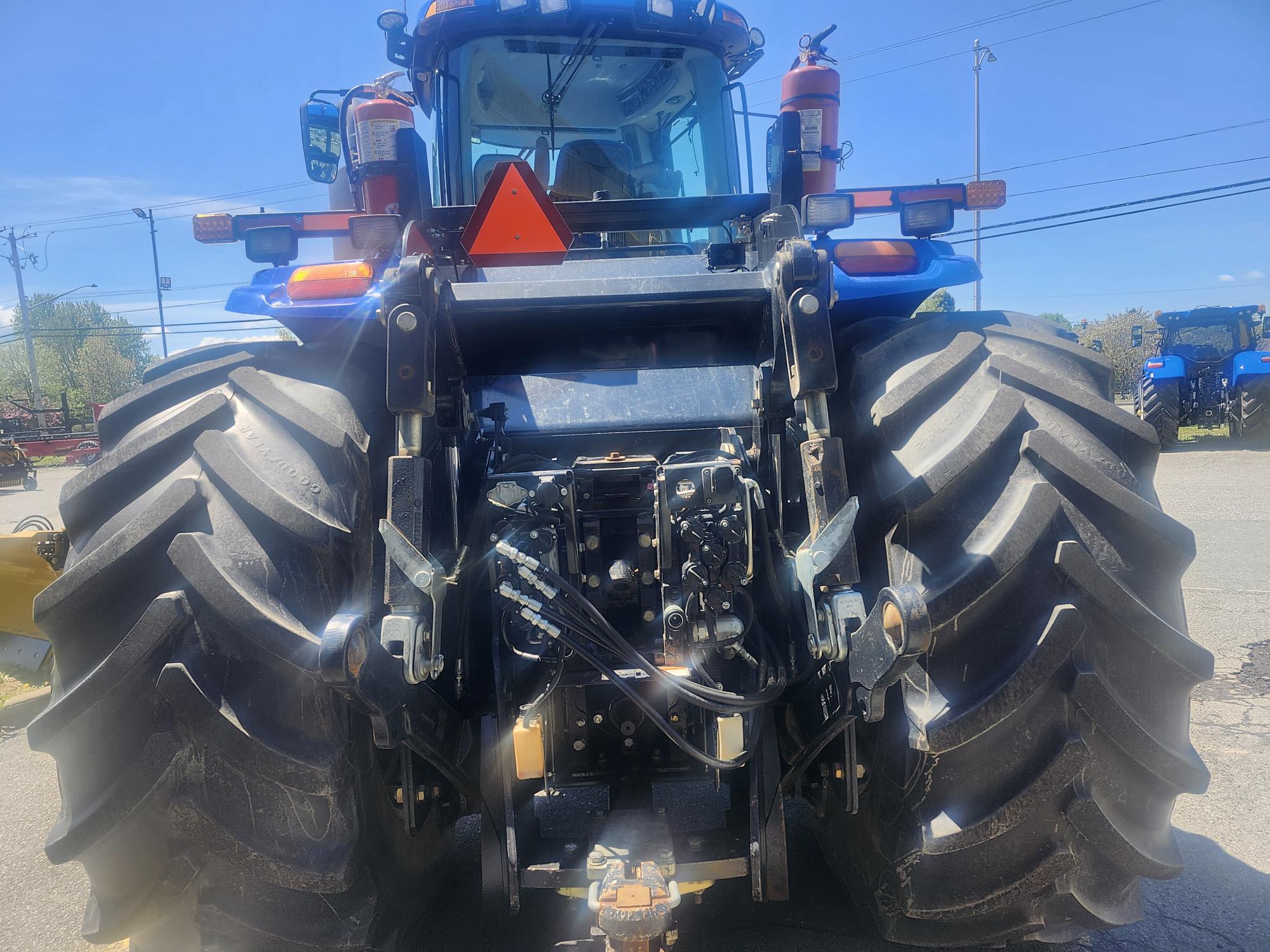 Vue arrière d'un tracteur bleu avec de gros pneus noirs, des raccords hydrauliques et deux extincteurs montés à l'arrière. Le ciel est dégagé et d'autres tracteurs sont visibles en arrière-plan.