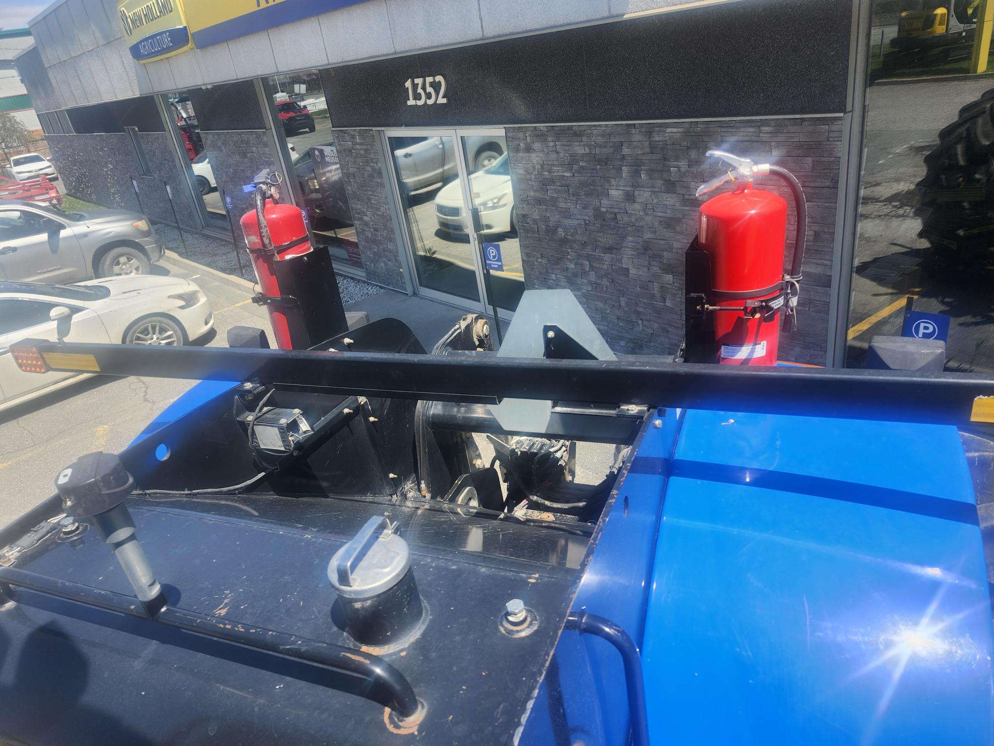 Vue de dessus d'un véhicule industriel bleu montrant deux gros extincteurs rouges montés à l'arrière, garé devant un bâtiment avec des vitres réfléchissantes et le numéro 1352 visible.