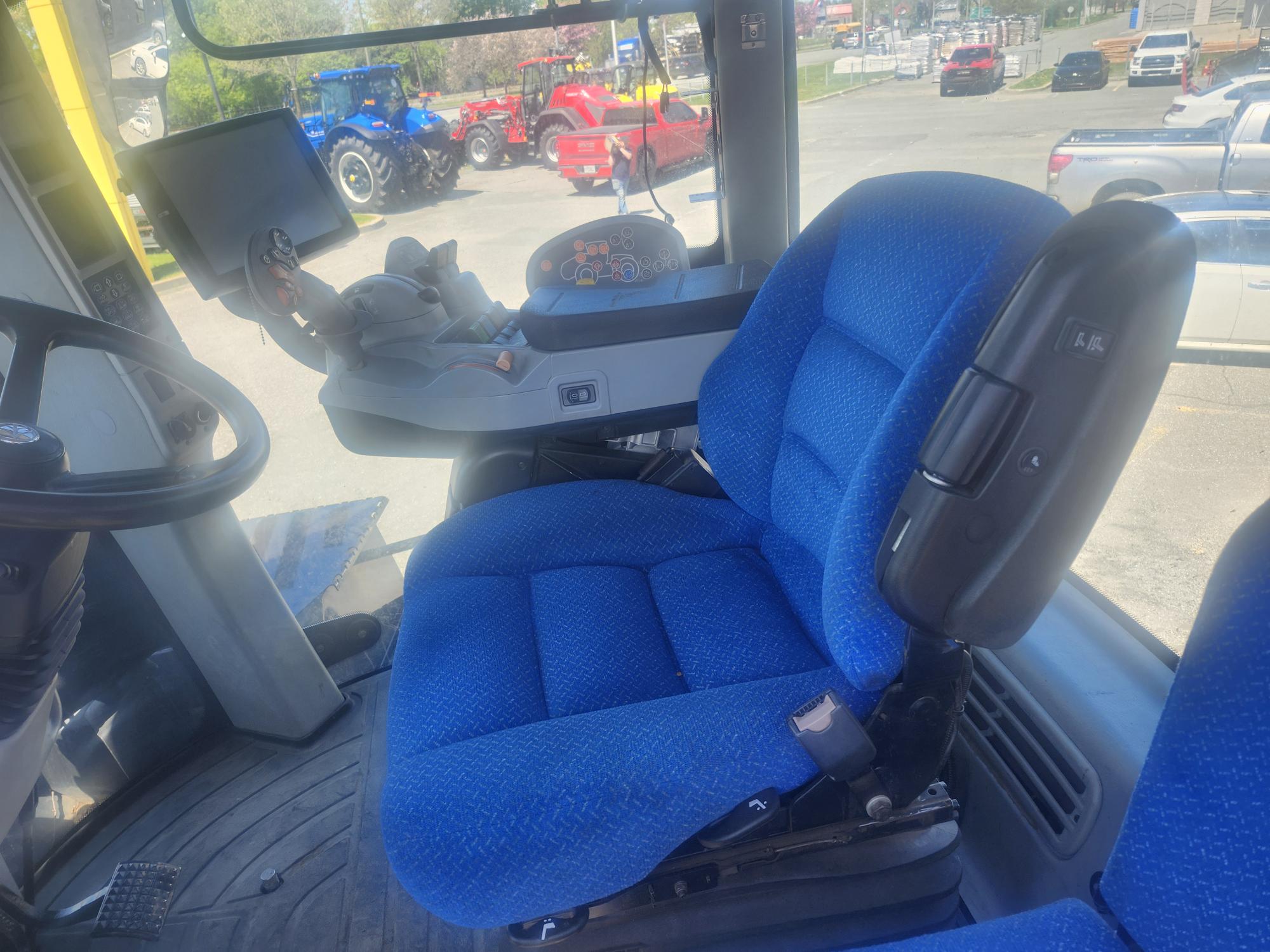 Siège conducteur et panneau de commande à l'intérieur d'une cabine de tracteur bleu moderne, avec volant et commandes électroniques visibles. Derrière les fenêtres, voitures et tracteurs sont garés sur un parking par une journée ensoleillée.