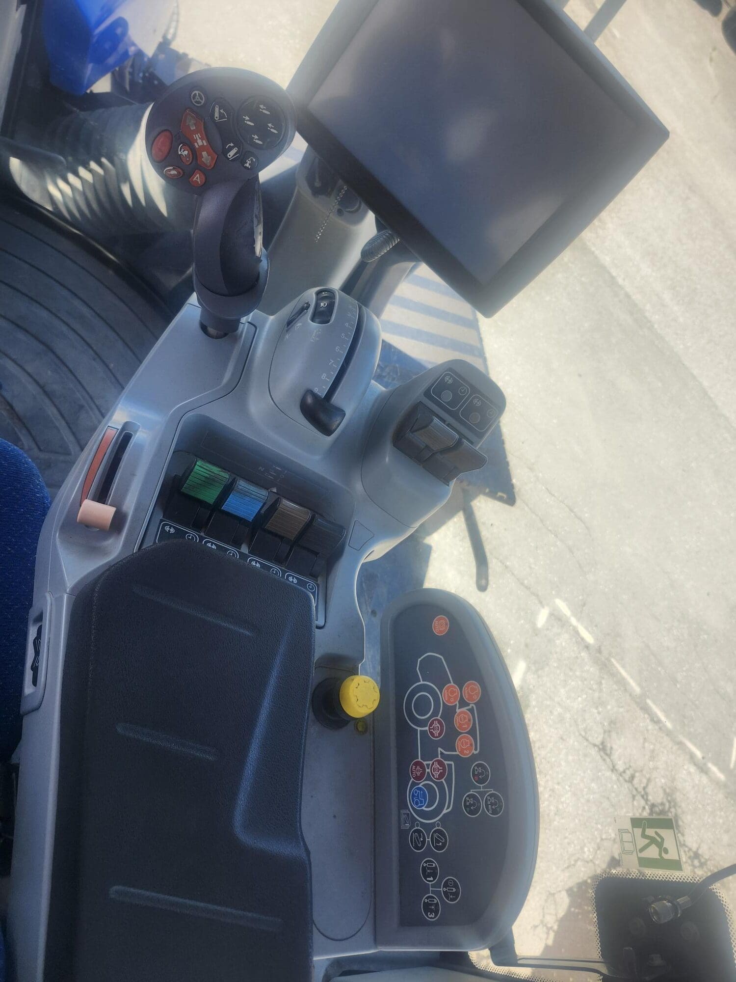 Gros plan d'un panneau de commande moderne pour tracteur ou machine, doté d'un écran tactile, de boutons de couleur, de leviers, d'un joystick et de divers interrupteurs. Le sol et une partie d'un siège sont visibles.