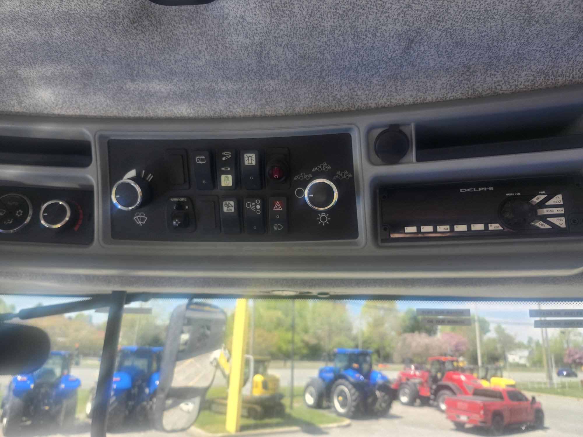 Panneau de commande à l'intérieur de la cabine d'un véhicule, affichant divers boutons, interrupteurs et une radio. À l'extérieur du pare-brise, plusieurs tracteurs colorés sont garés sur un parking, avec des arbres et une clôture en arrière-plan.