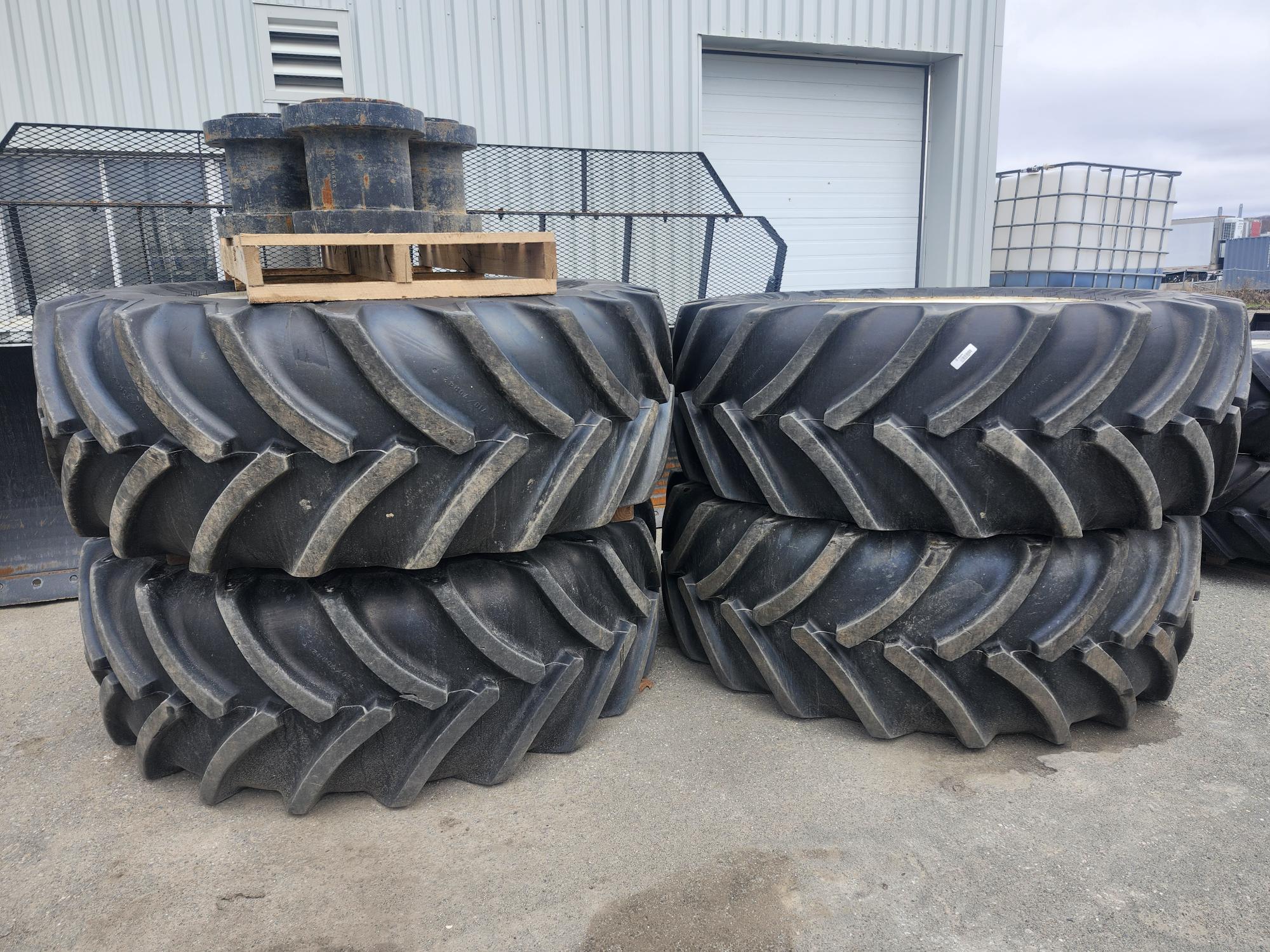 Quatre gros pneus de tracteur robustes sont empilés en deux piles à l'extérieur d'un bâtiment métallique, surmontés d'une palette en bois et d'un objet métallique. Le sol est en gravier et des équipements industriels apparaissent en arrière-plan.