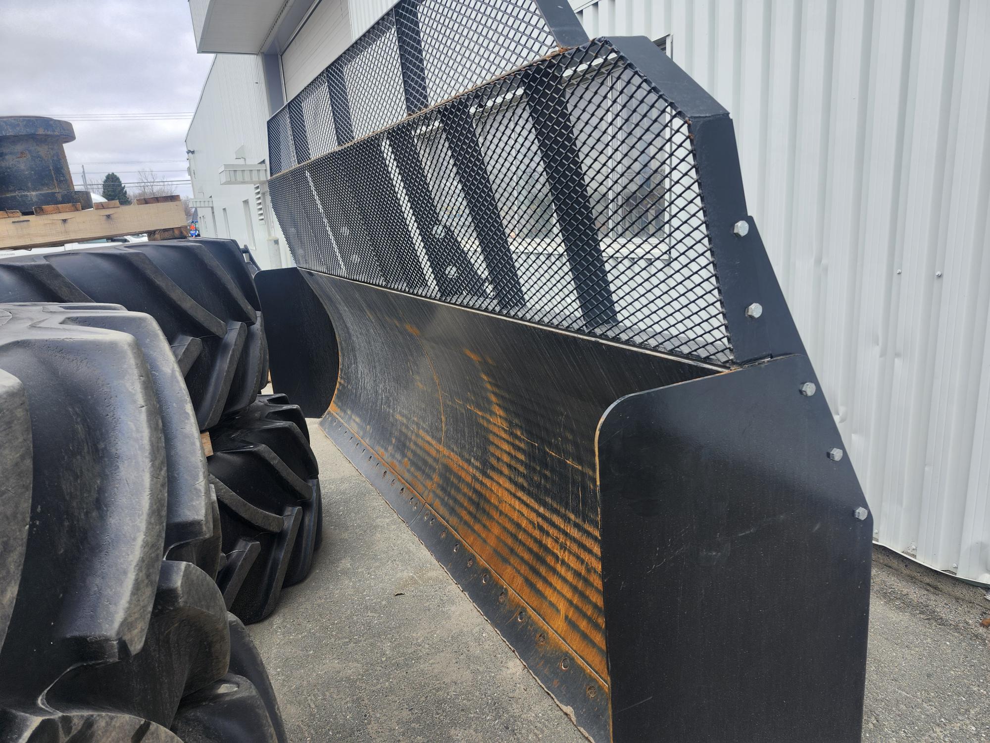 Une grande lame métallique industrielle avec un grillage de protection, positionnée verticalement à côté de gros pneus de tracteur à l'extérieur d'un bâtiment en tôle ondulée.