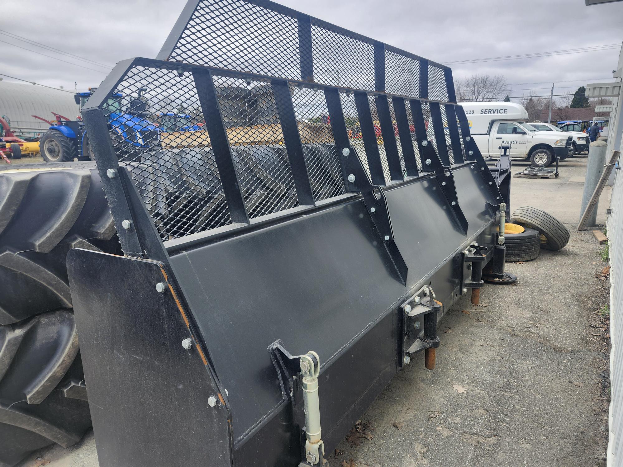Un grand accessoire de chargeuse compacte en métal avec une protection grillagée et des lames noires est exposé à l'extérieur sur un terrain pavé, entouré de pneus, de camions et d'autres machines.