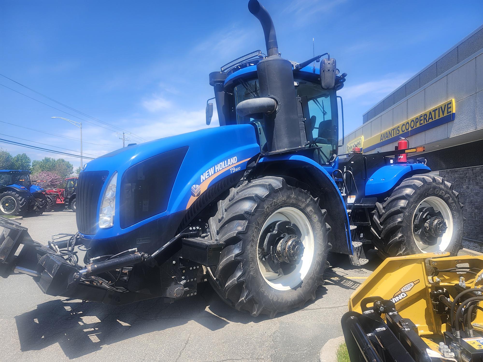 Un gros tracteur New Holland bleu est garé devant un bâtiment arborant un panneau jaune indiquant « Adams Cooperative » par une journée ensoleillée. Le tracteur est équipé de gros pneus et de matériel agricole.