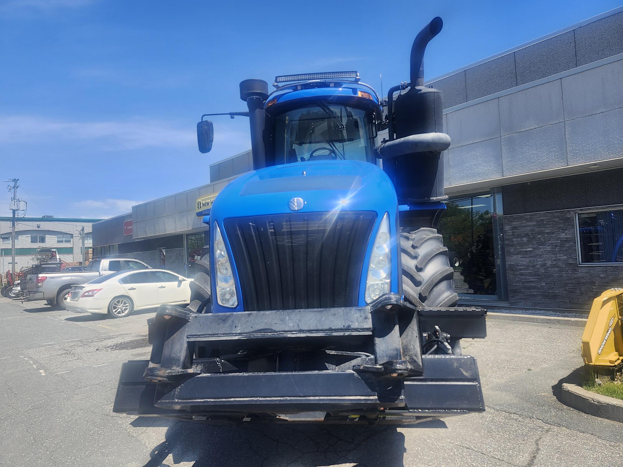 Un gros tracteur bleu garé devant un bâtiment commercial, avec plusieurs voitures blanches garées à proximité par une journée ensoleillée.