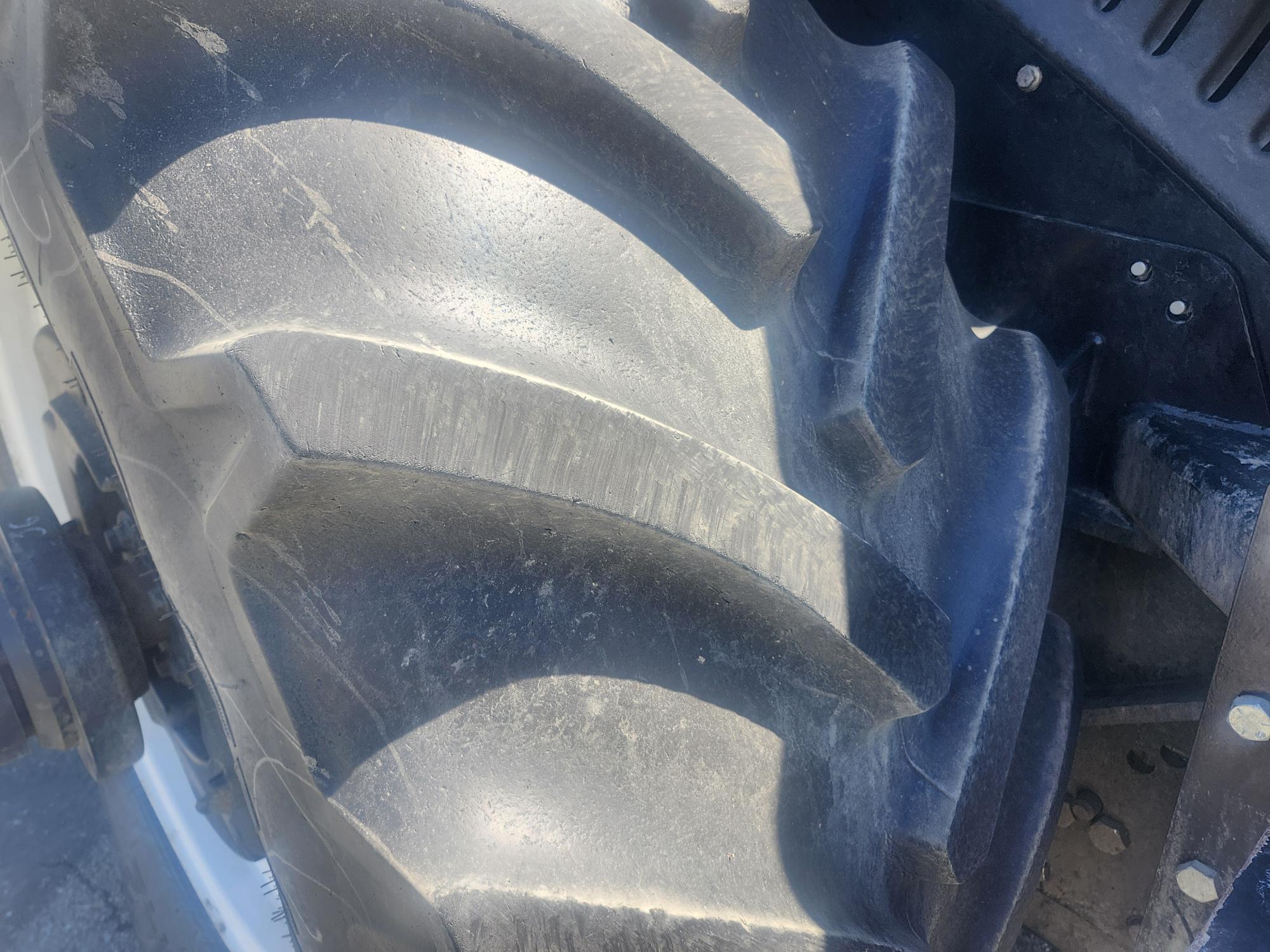 Gros plan d'un gros pneu de tracteur robuste aux sculptures profondes et inclinées. Le pneu semble usé et poussiéreux, et une partie de la roue métallique et de la machinerie est visible à droite.