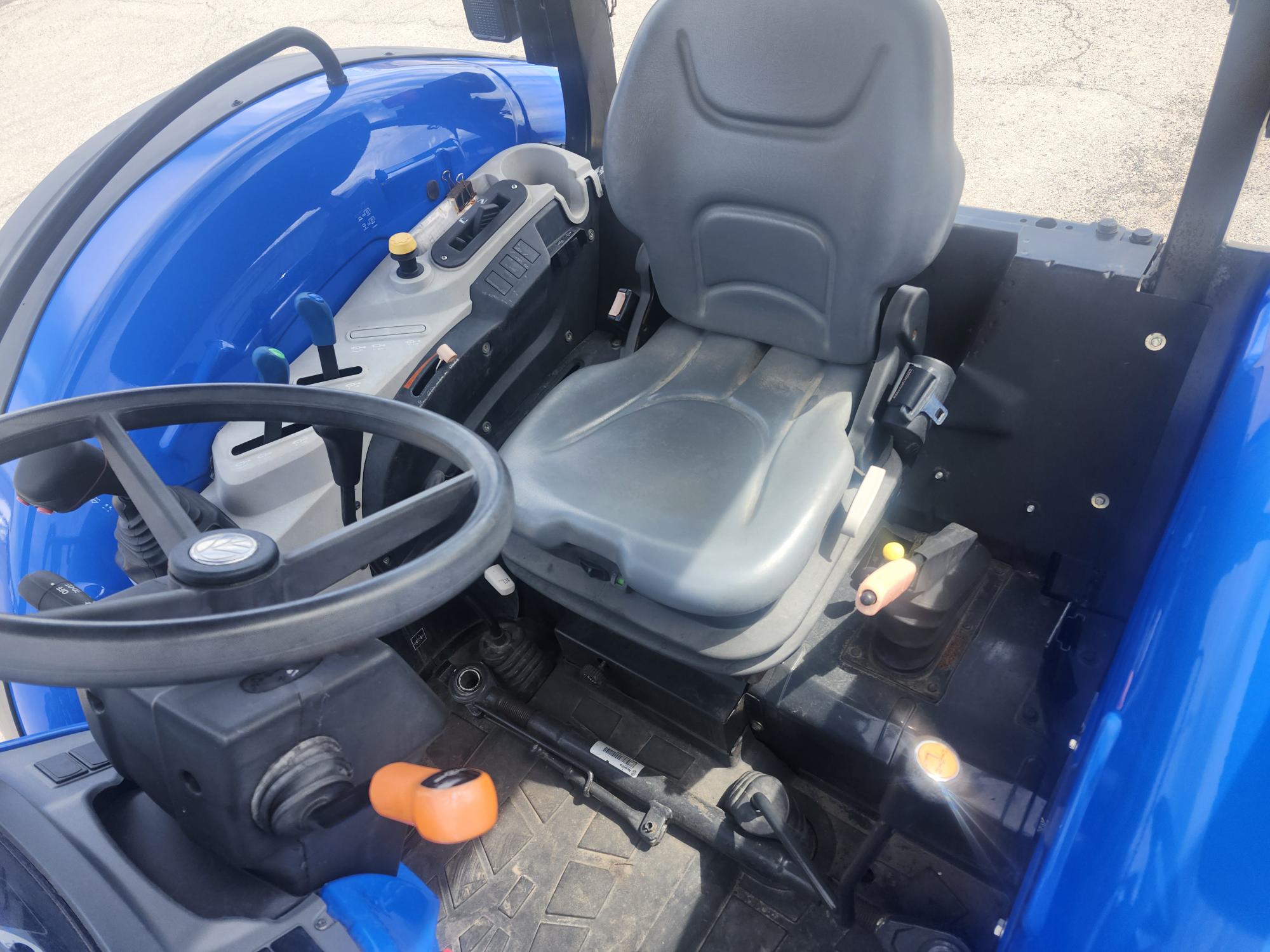 Vue rapprochée du siège conducteur et des commandes d'un tracteur bleu, montrant le volant, les leviers de vitesses, les pédales et un siège gris rembourré avec accoudoirs par une journée ensoleillée.