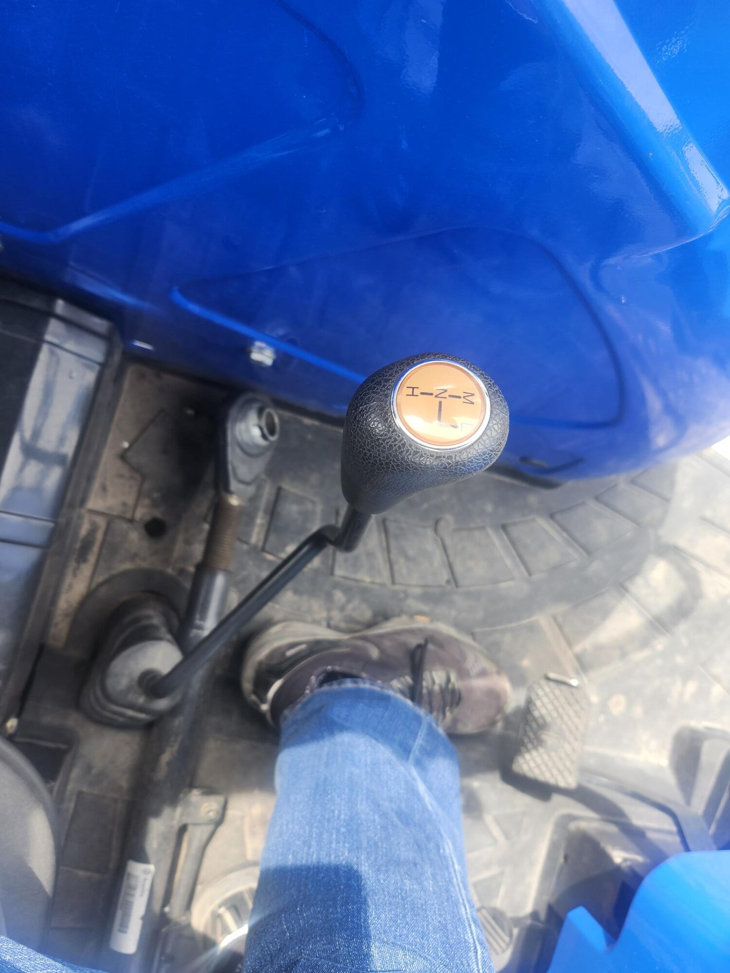 Une personne portant un jean et des bottes actionne un levier de vitesse manuel à l'intérieur d'un véhicule bleu, probablement un tracteur. Le pommeau du levier de vitesse affiche un schéma de vitesses simple.