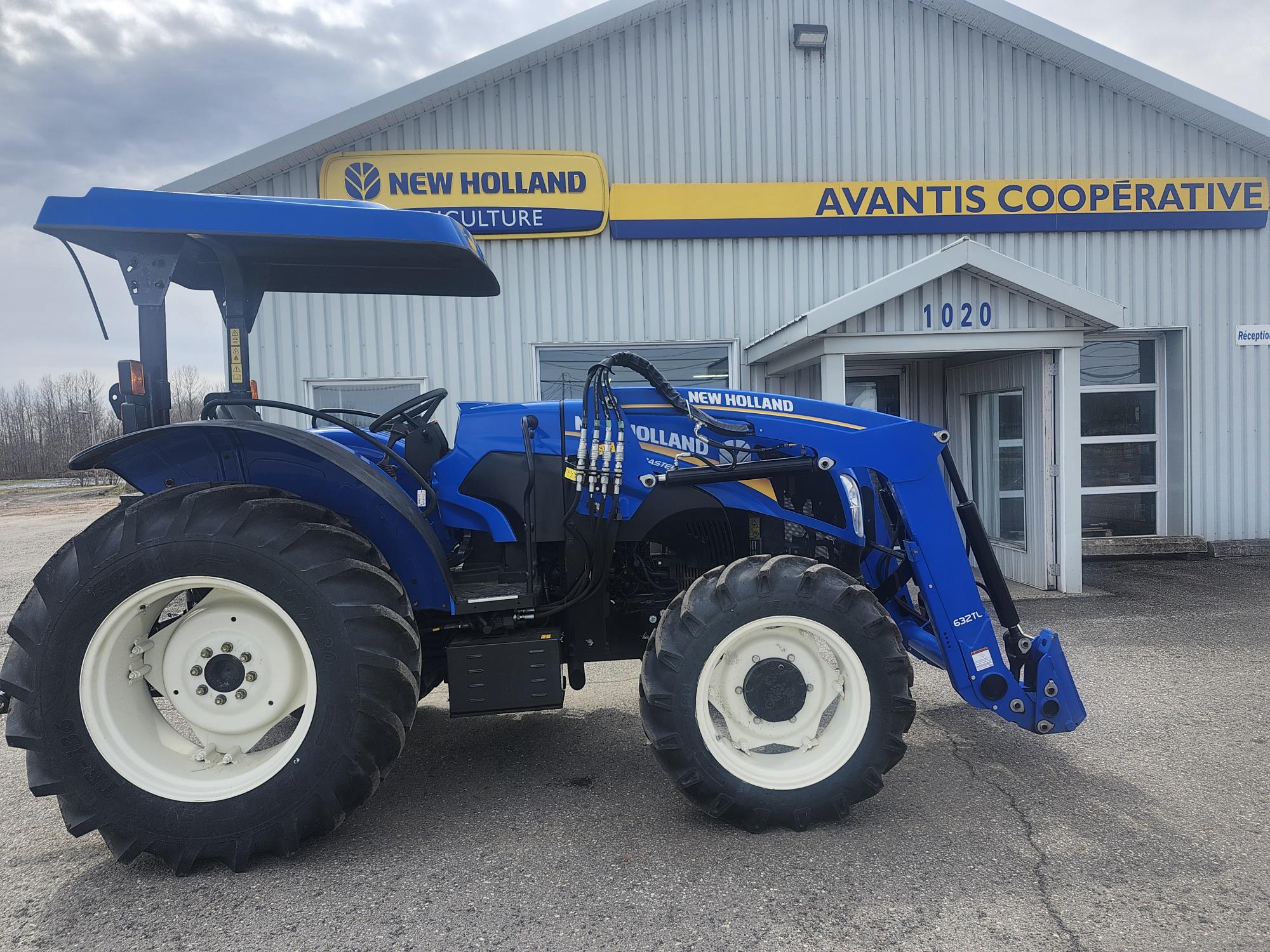 Un tracteur New Holland bleu est garé devant un bâtiment portant l'inscription « Avantis Cooperative » avec un panneau « New Holland Agriculture » au-dessus de l'entrée.