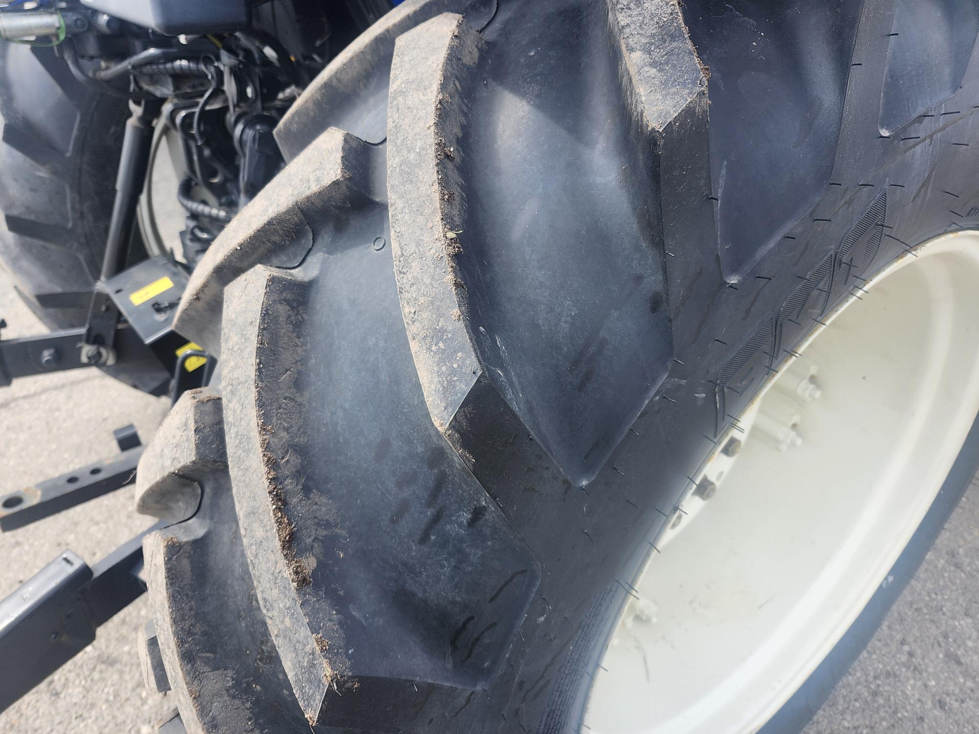 Gros plan d'un gros pneu de tracteur noir à sculptures profondes, monté sur une jante blanche. L'arrière-plan montre une partie du tracteur et une route goudronnée.