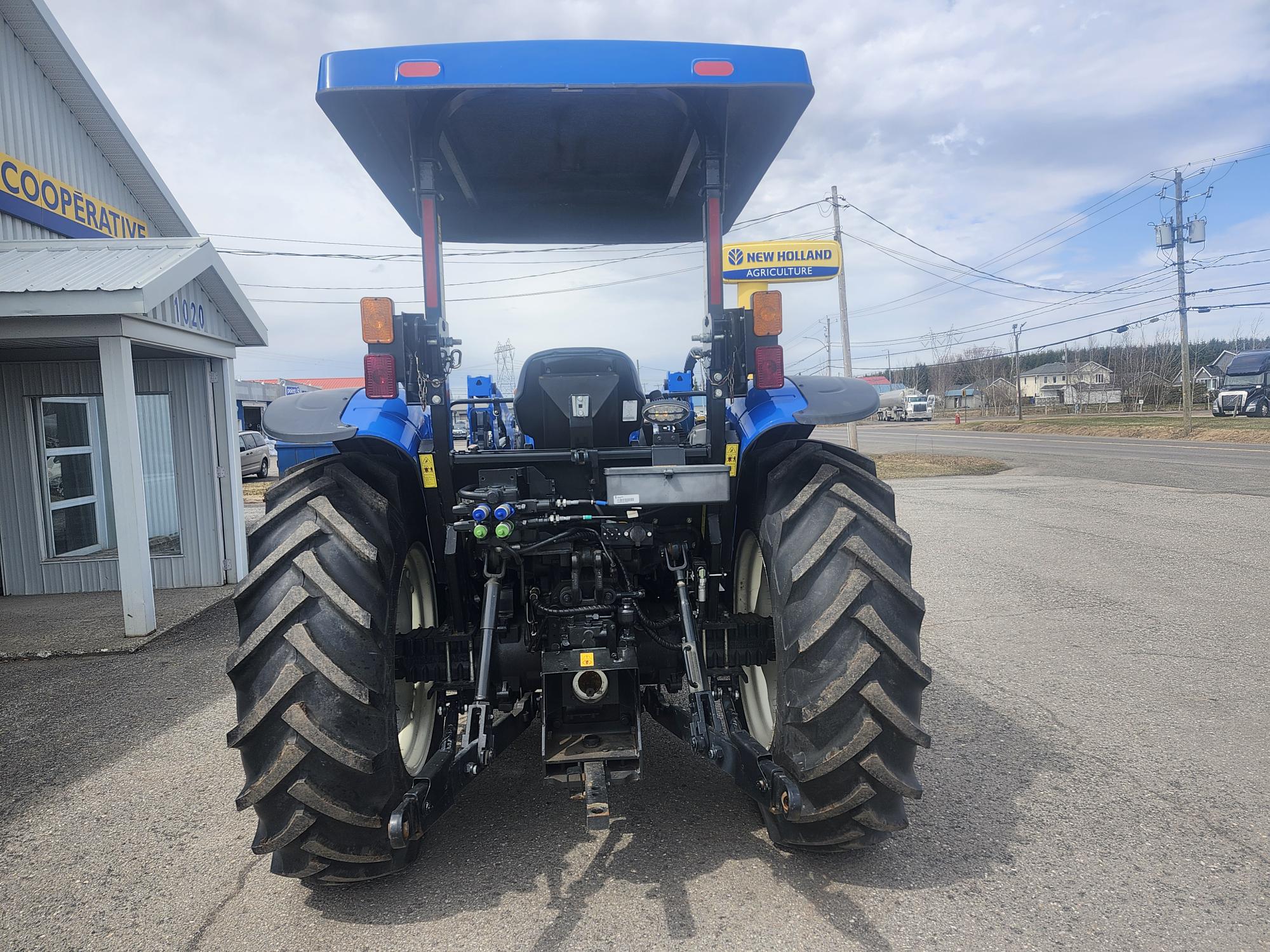 Un tracteur bleu vu de derrière est garé sur un terrain pavé près d'un bâtiment et d'un panneau de concessionnaire New Holland, avec un ciel nuageux et des lignes électriques en arrière-plan.