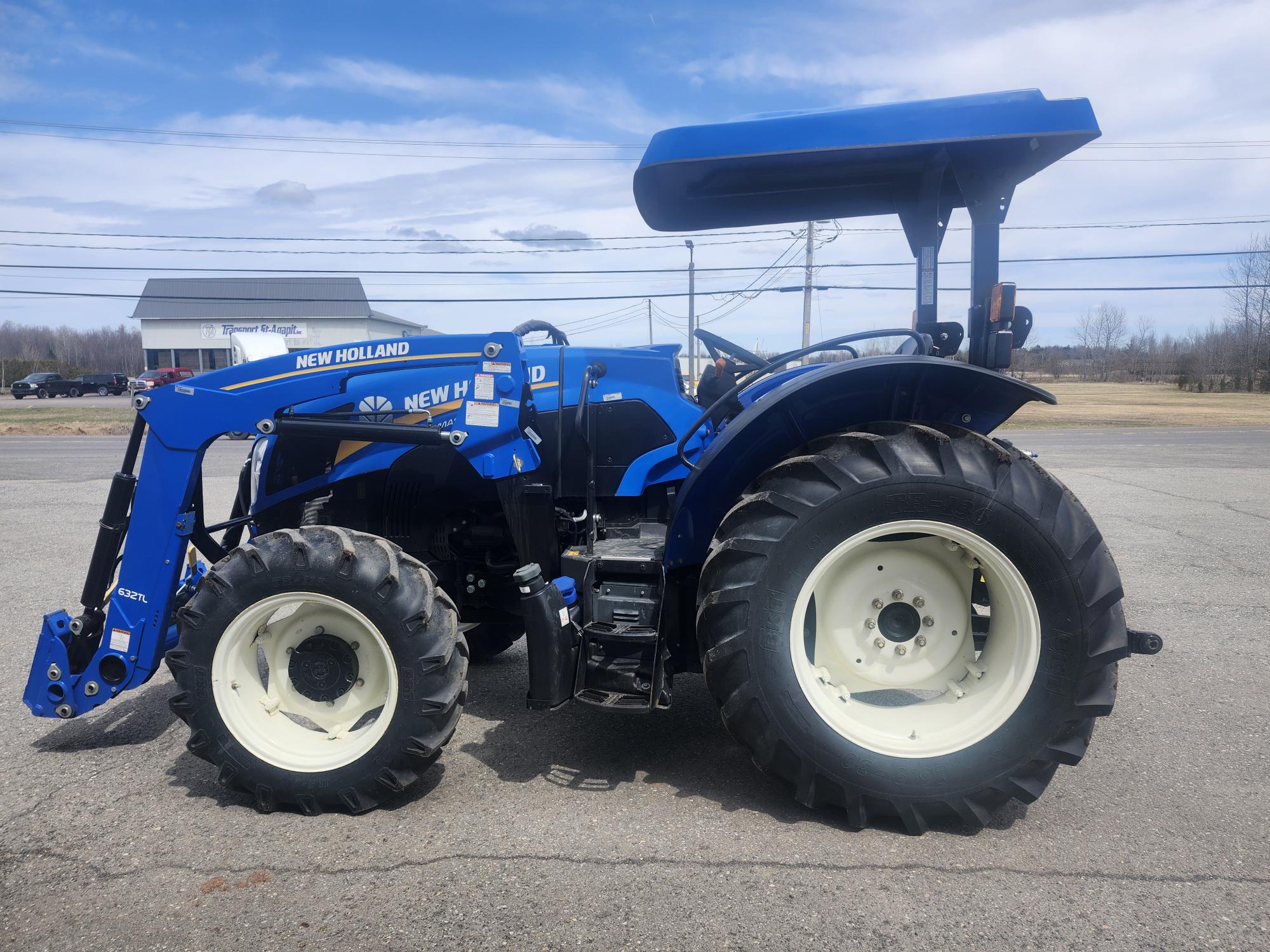 Un tracteur New Holland bleu équipé d'un chargeur frontal et d'un toit ouvrant est garé sur une surface pavée, avec un champ et un bâtiment en arrière-plan sous un ciel partiellement nuageux.