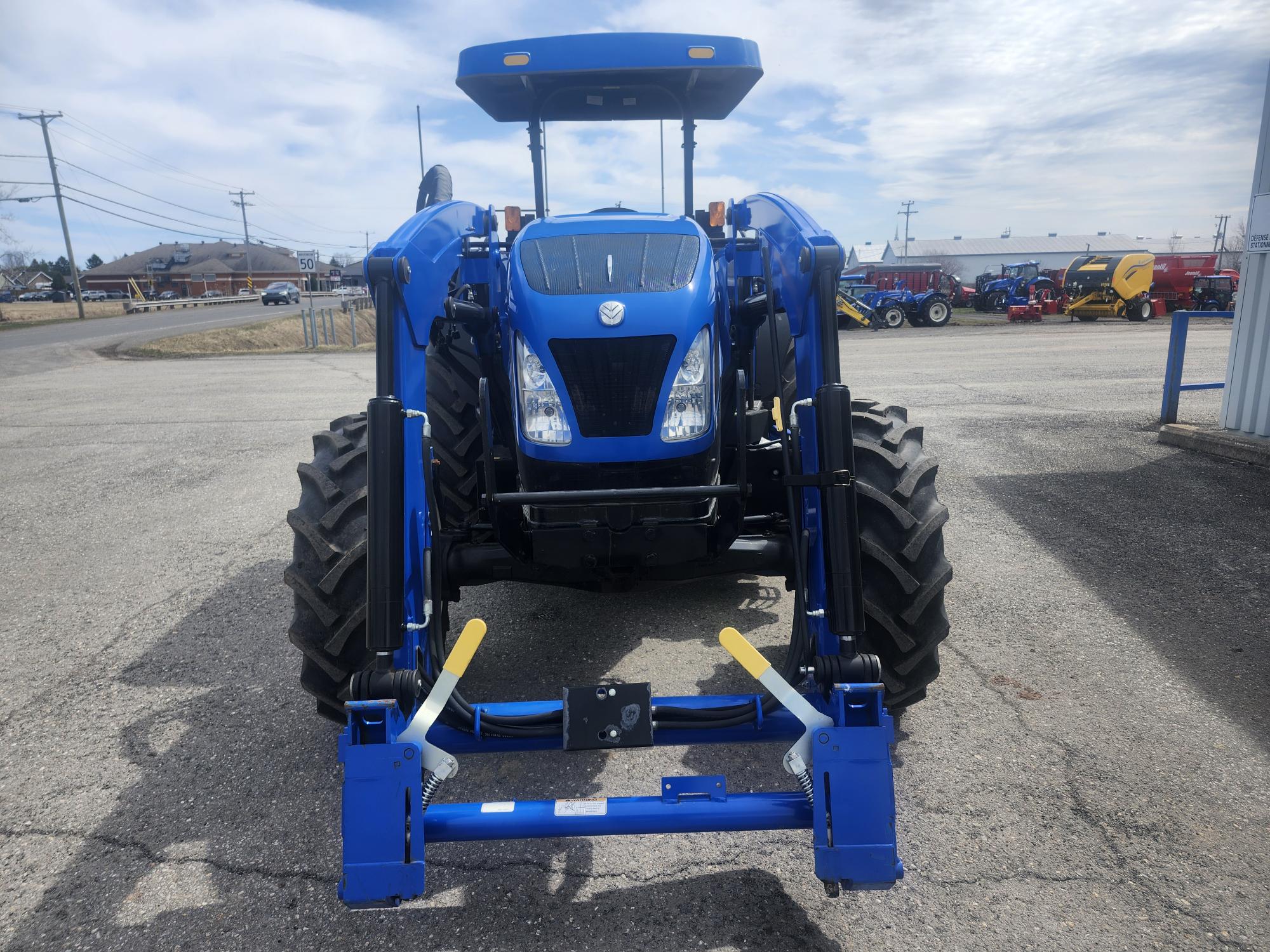 Un tracteur bleu avec de gros pneus noirs est garé sur un terrain pavé, face à la route. Il est équipé d'un chargeur frontal et est entouré d'autres équipements agricoles en arrière-plan, sous un ciel partiellement nuageux.