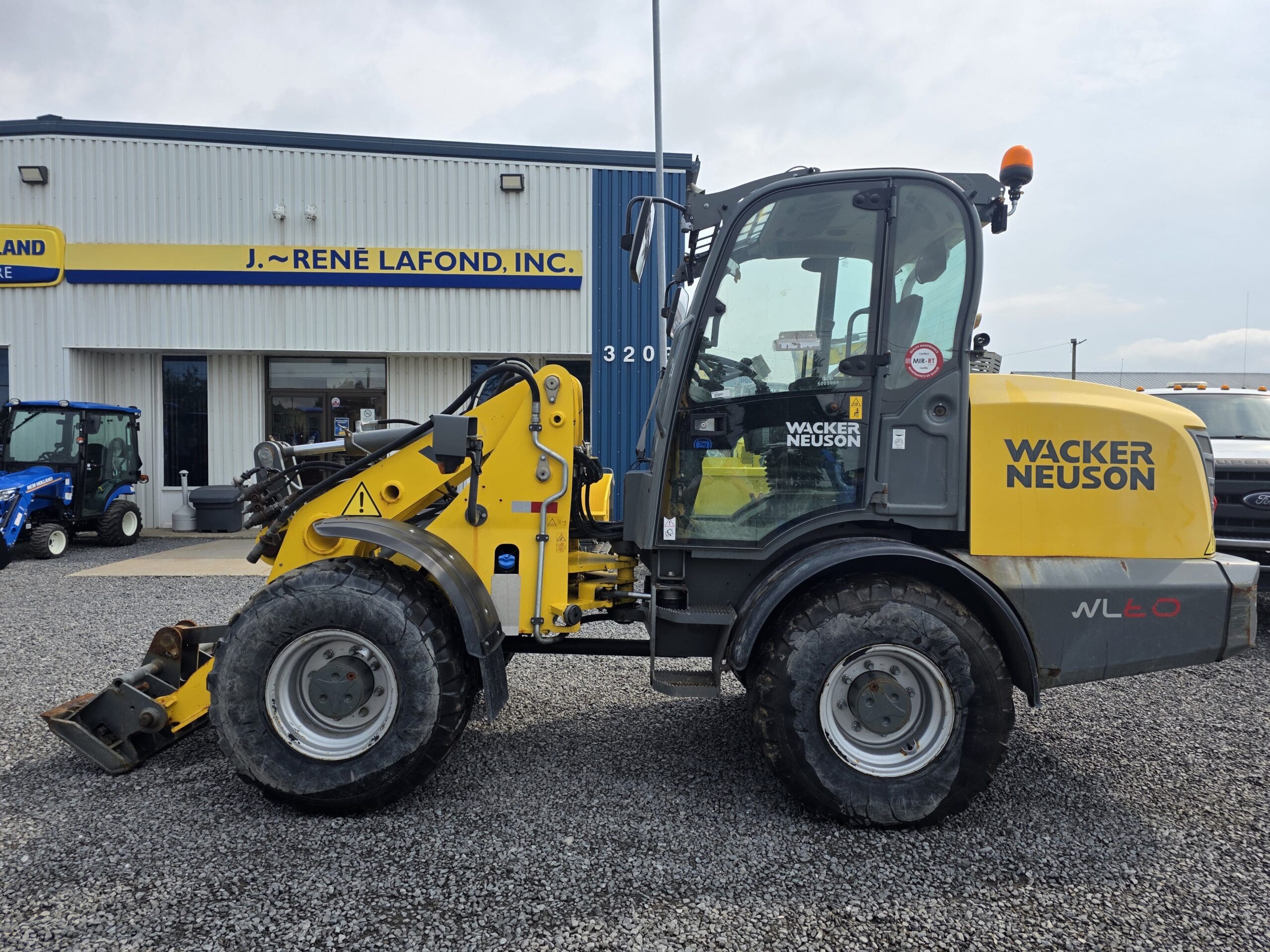 Une chargeuse sur pneus Wacker Neuson jaune est garée sur du gravier devant un bâtiment portant l'inscription