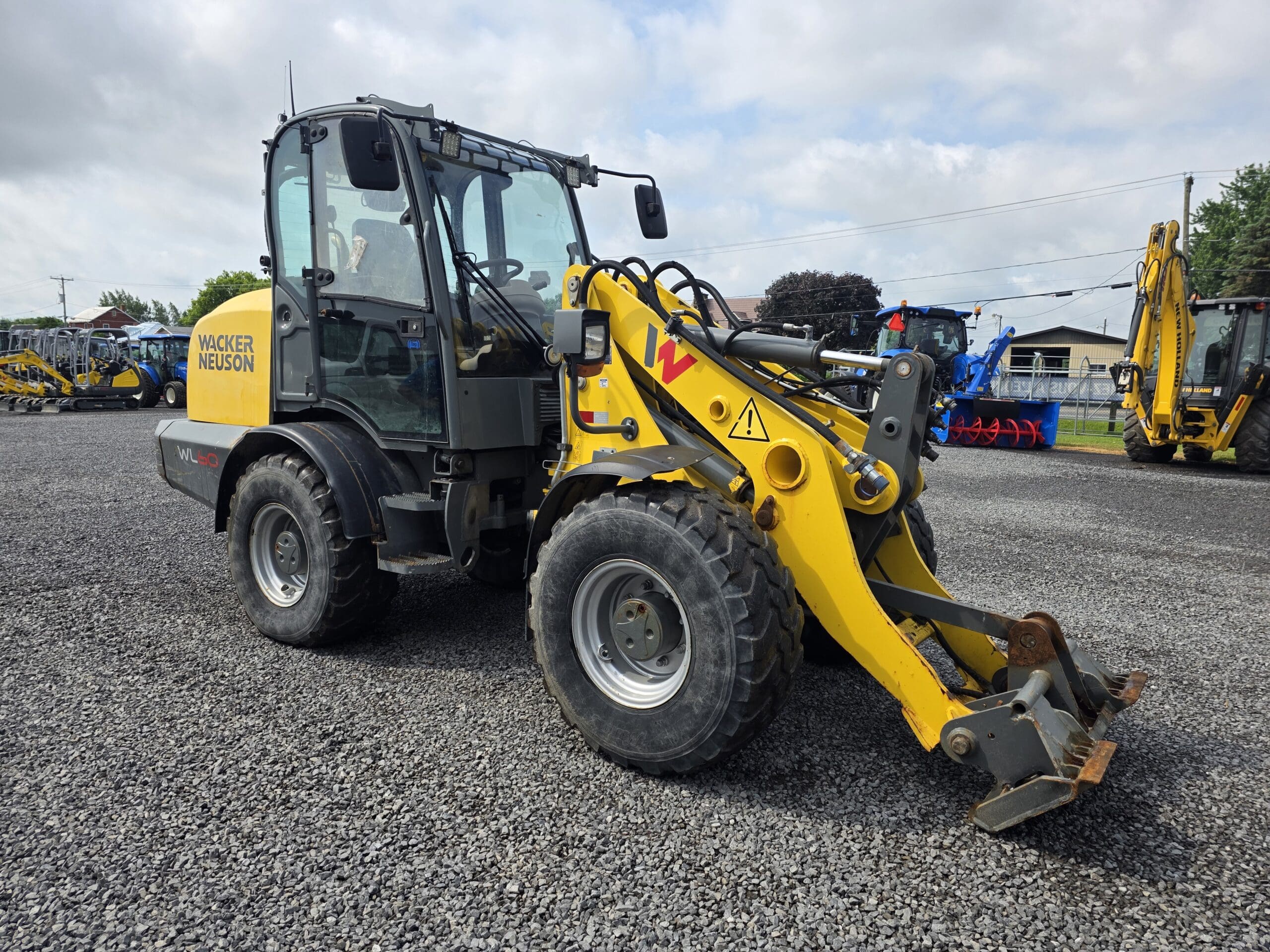 Une chargeuse sur pneus Wacker Neuson jaune avec un accessoire frontal est garée sur un terrain en gravier avec d'autres équipements de construction visibles à l'arrière-plan sous un ciel nuageux.